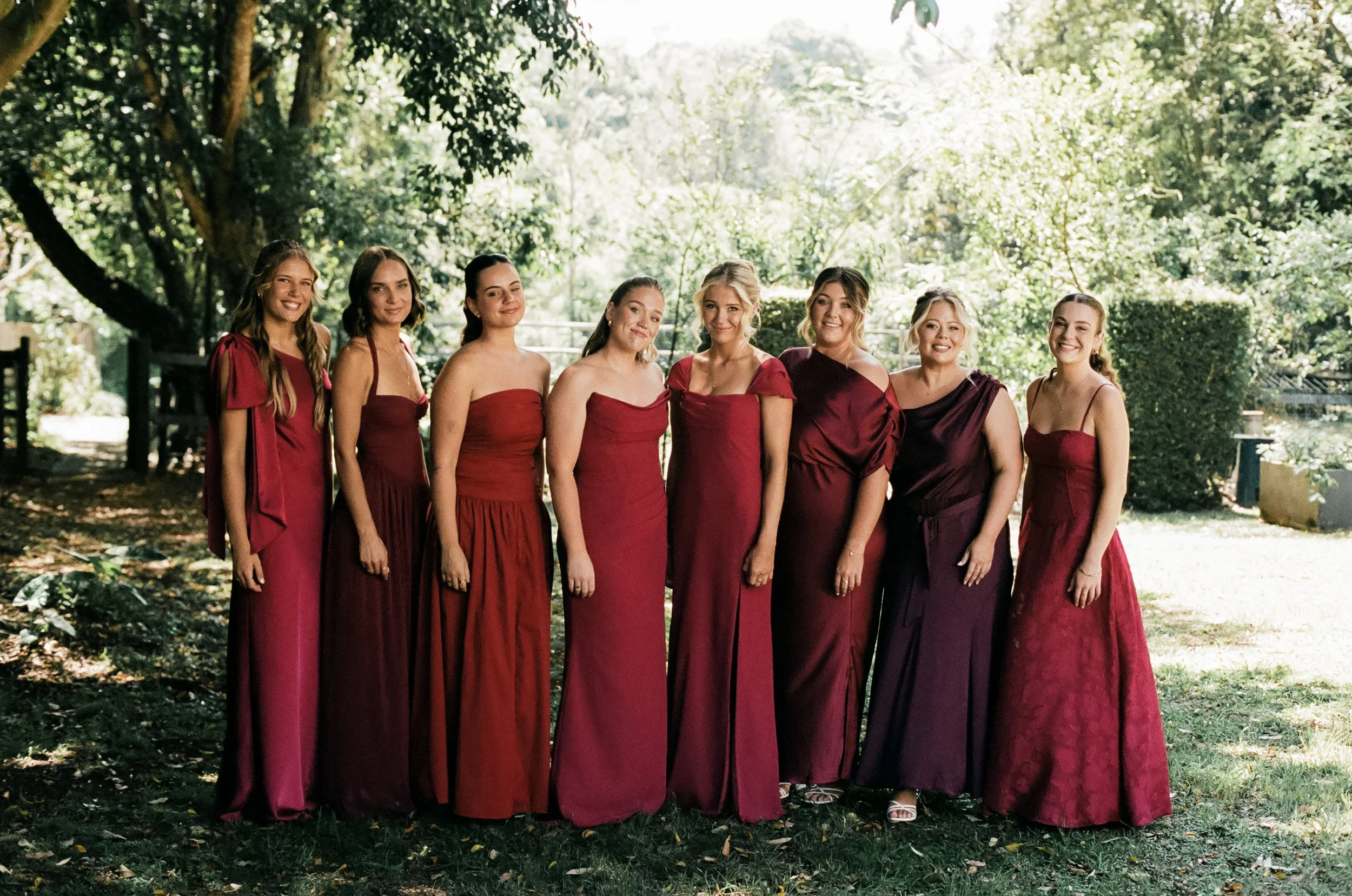 A group of nine women standing outdoors on grass, dressed in various shades of red and burgundy dresses, smiling for a photo in a sunny, wooded area.