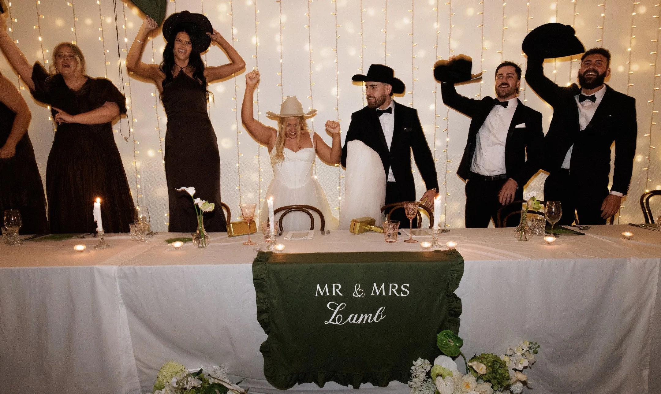 A group of joyous people at a wedding reception standing behind a table decorated with candles and floral arrangements, celebrating the newlywed couple with hats.