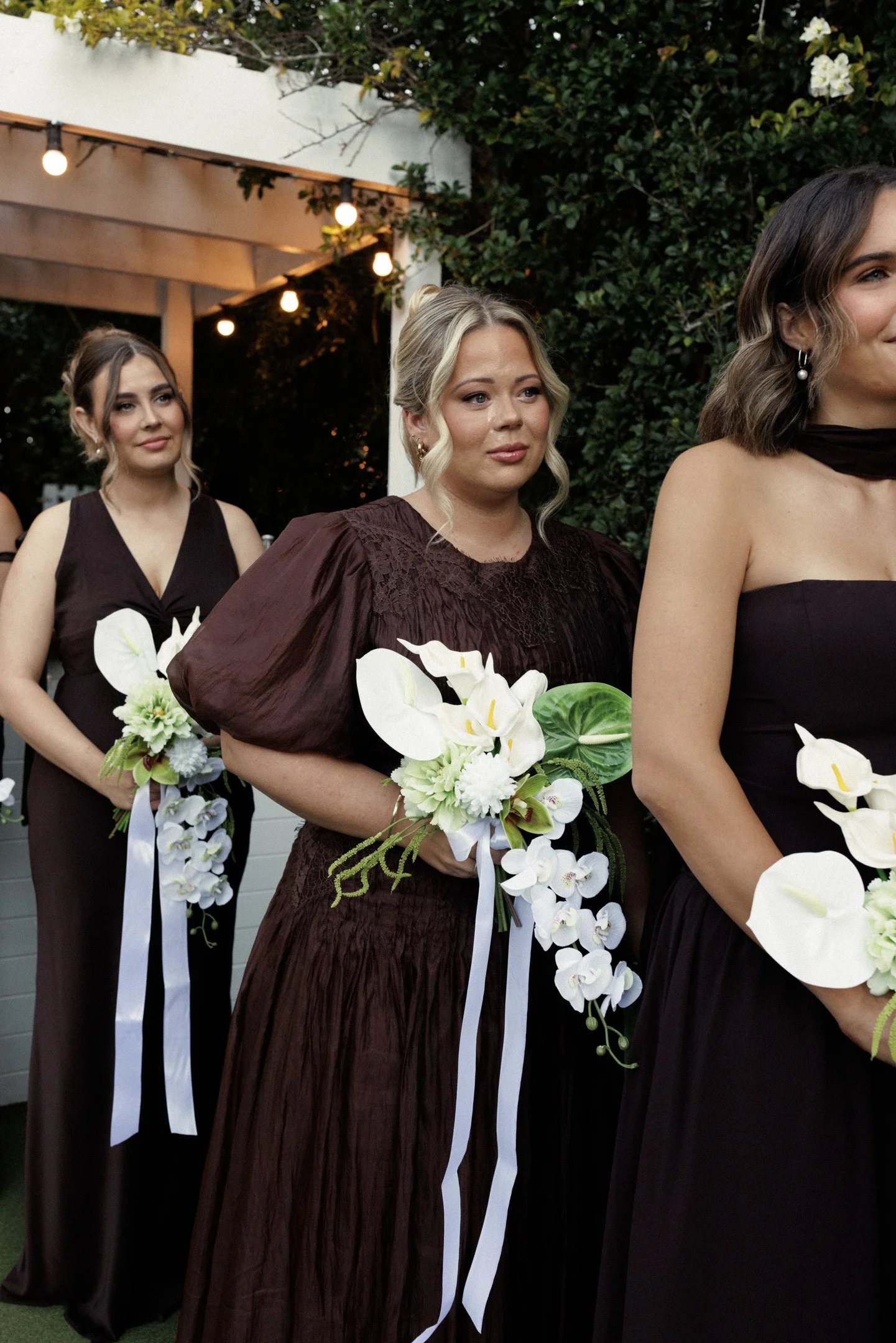 Women dressed in formal dark dresses holding white floral bouquets at an outdoor event.
