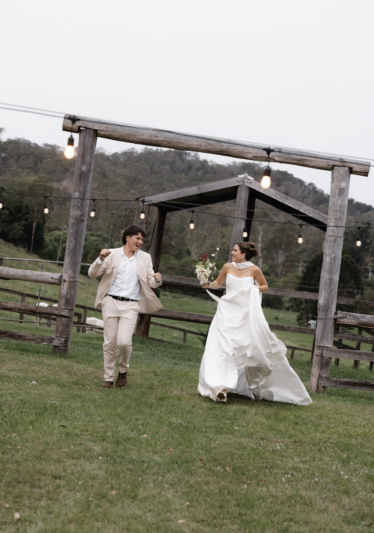 A bride and groom celebrating outdoors on their wedding day, with the bride holding a bouquet and the groom expressing joy, surrounded by a rustic wooden fence, string lights, and scenic hilly landscape in the background.