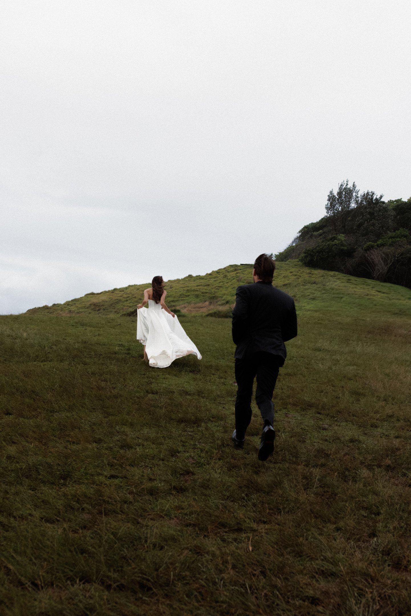 A woman in a white dress running up a grassy hill with a man in a black suit behind her, on a cloudy day