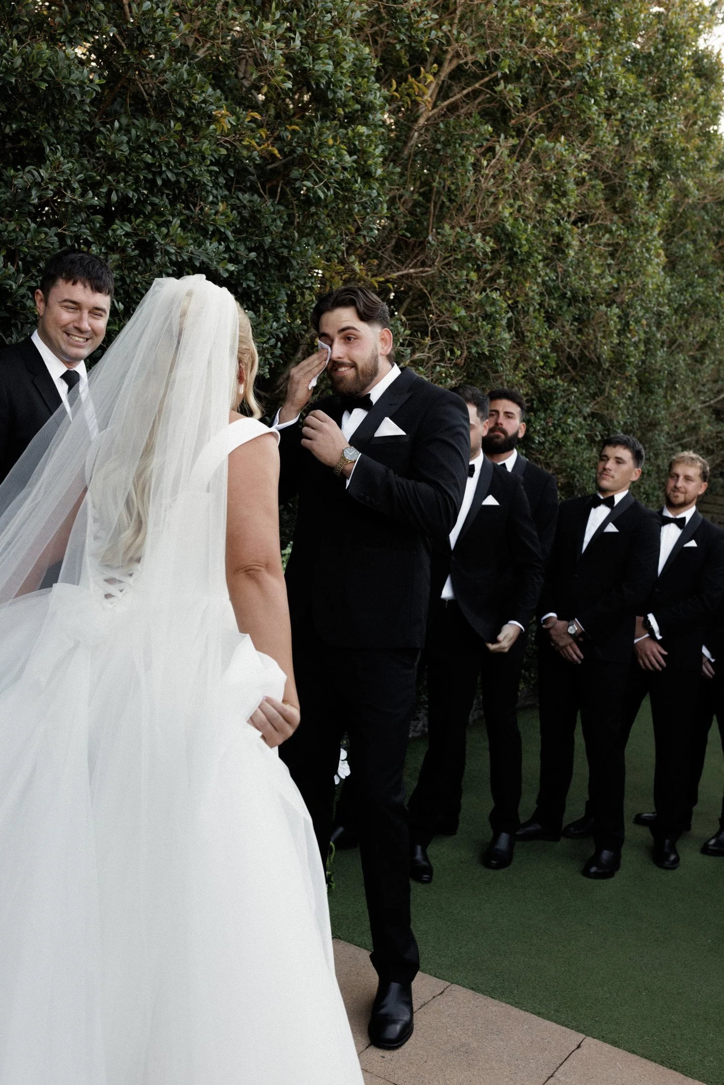 A wedding ceremony outdoors with a bride, groom, and groomsmen. The bride is wearing a wedding dress with a veil. The groom in a black tuxedo is wiping tears from his eyes, surrounded by groomsmen in tuxedos.