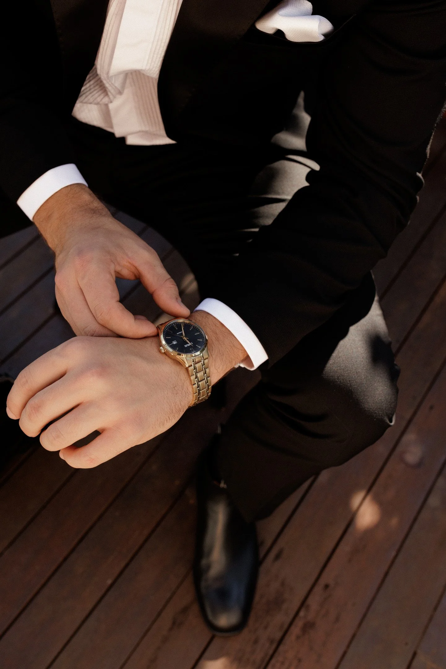Close-up of a person dressed in a black suit with white shirt cuffs, wearing a gold wristwatch, sitting at a wooden table, with another person's hand adjusting the watch.