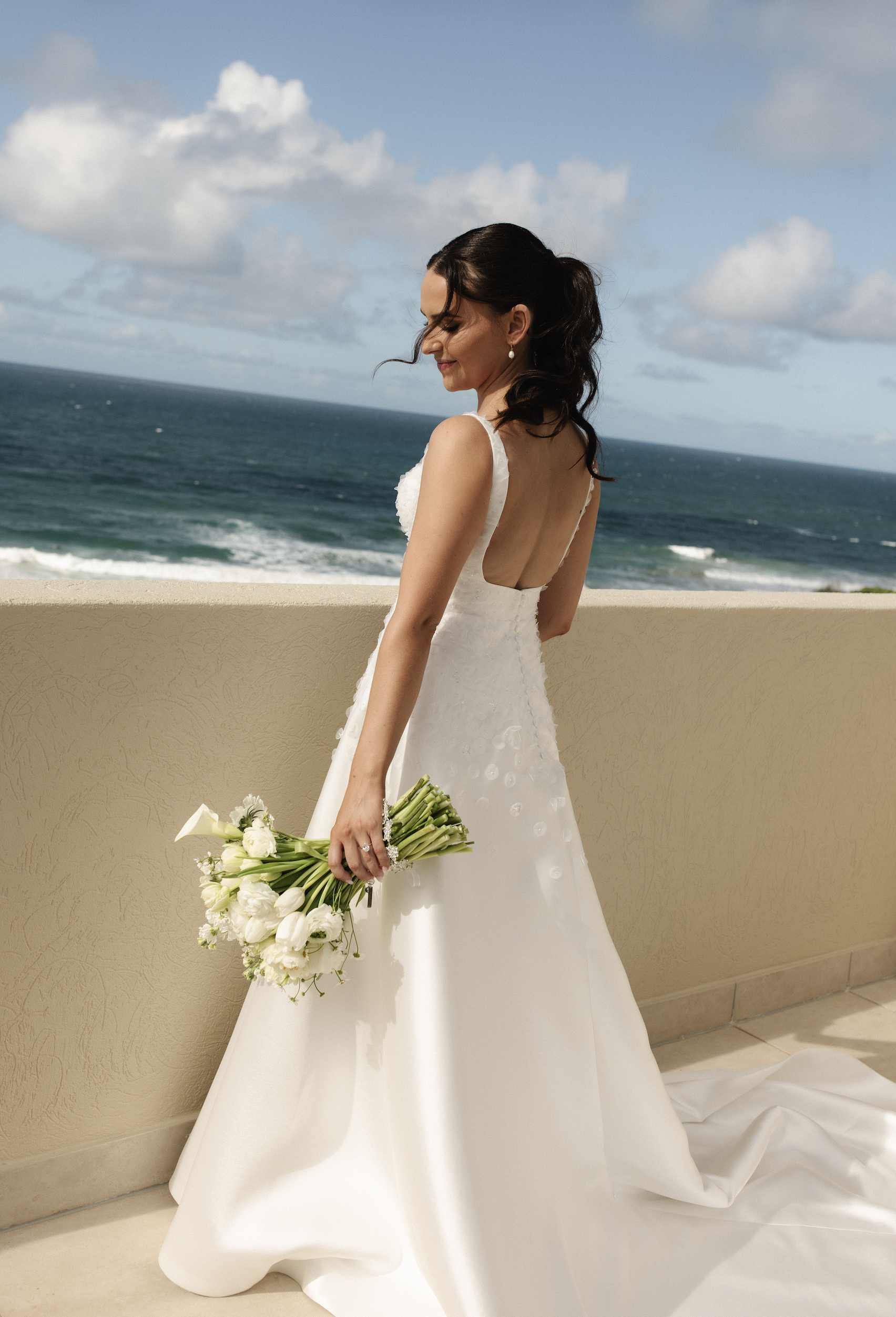 A bride in a white wedding dress holding a bouquet of white flowers, standing on a balcony overlooking the ocean with a partly cloudy sky.