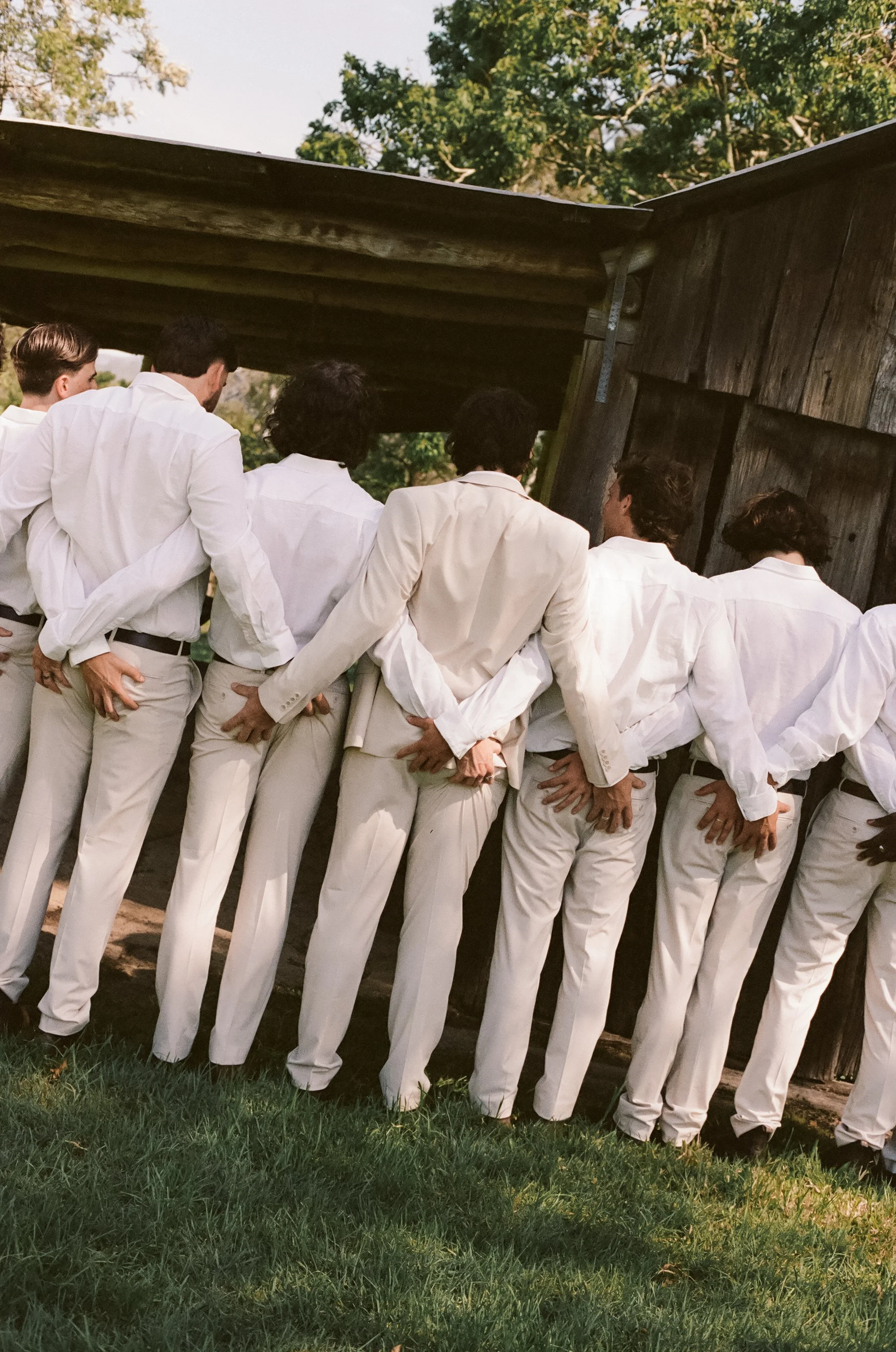 Group of men dressed in white suits standing in a row with their hands behind their backs, watching something through a wooden window during daytime.