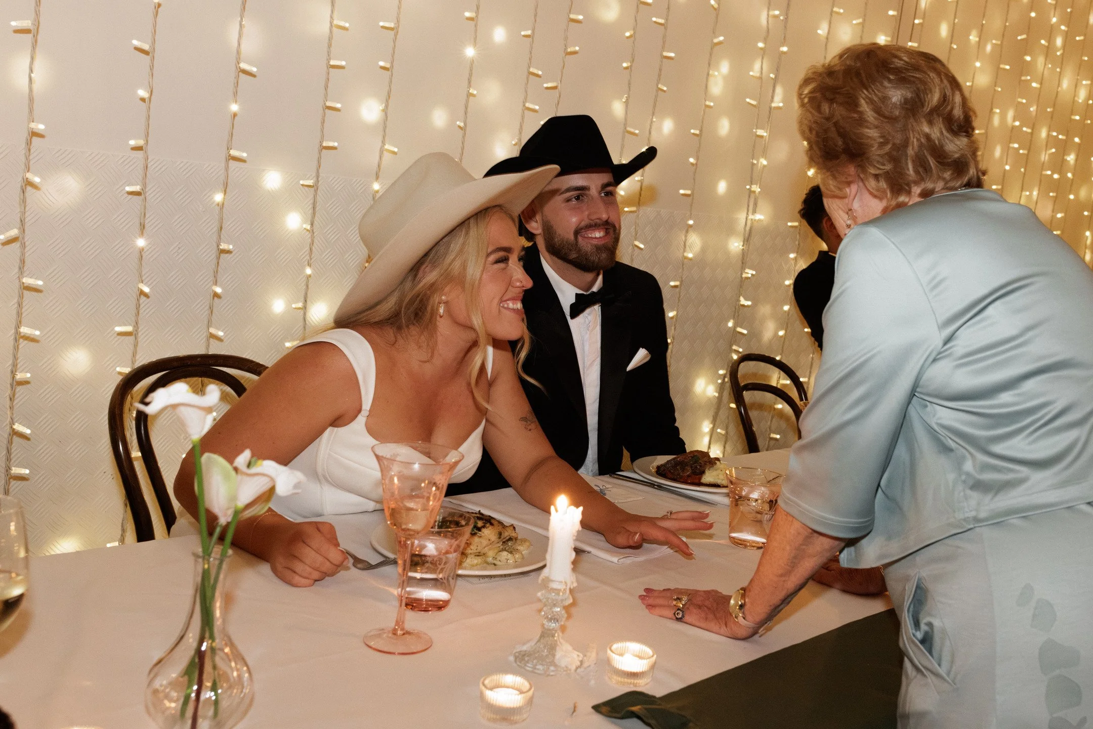 A woman with blonde hair wearing a wide-brimmed hat and a white dress is smiling at a man in a black tuxedo and cowboy hat. They are seated at a dinner table decorated with candles and flowers, in front of a backdrop of string lights. An older woman 