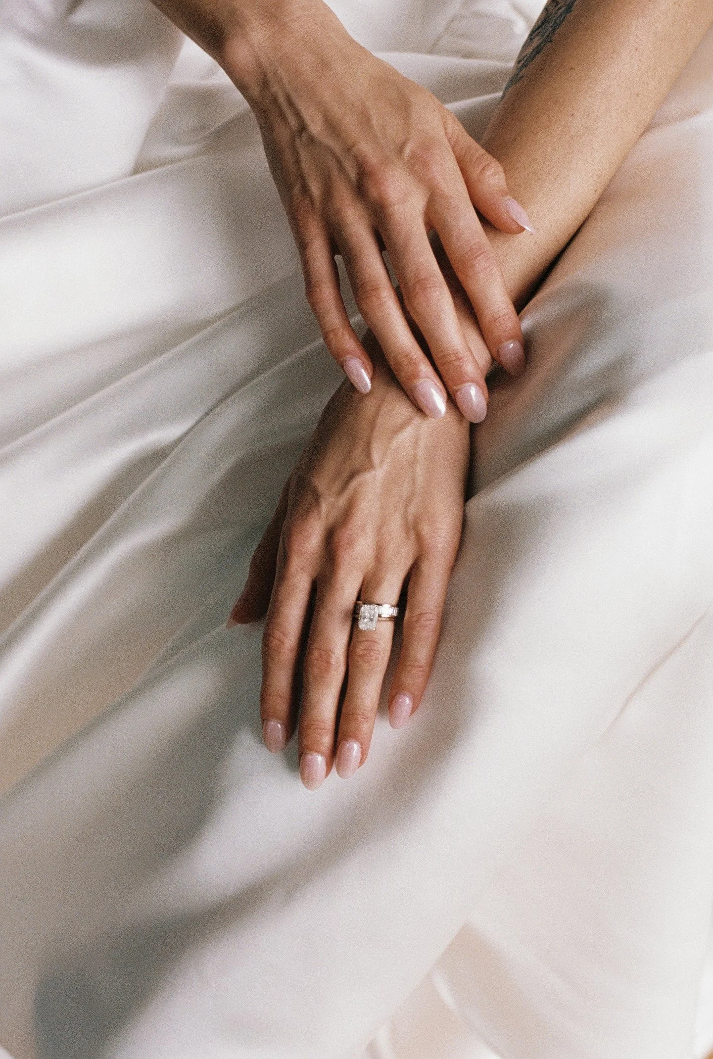 Two hands gently touching, one with a wedding ring, resting on a silky white fabric background.