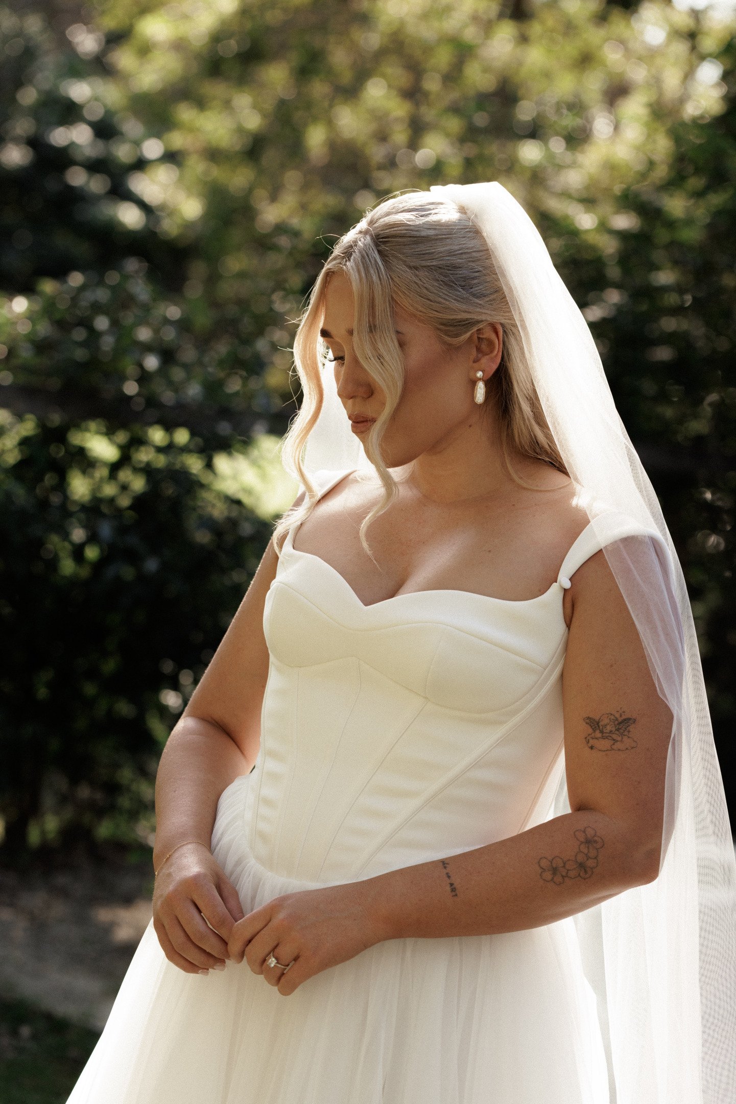 A bride in a white wedding dress standing outdoors with her eyes closed and her head slightly bowed, with natural sunlight and blurred green foliage in the background.