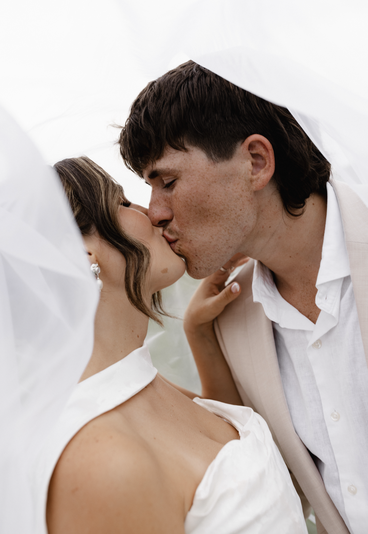A couple kissing under white fabric, with the woman at left and the man at right, both dressed in light-colored wedding attire.