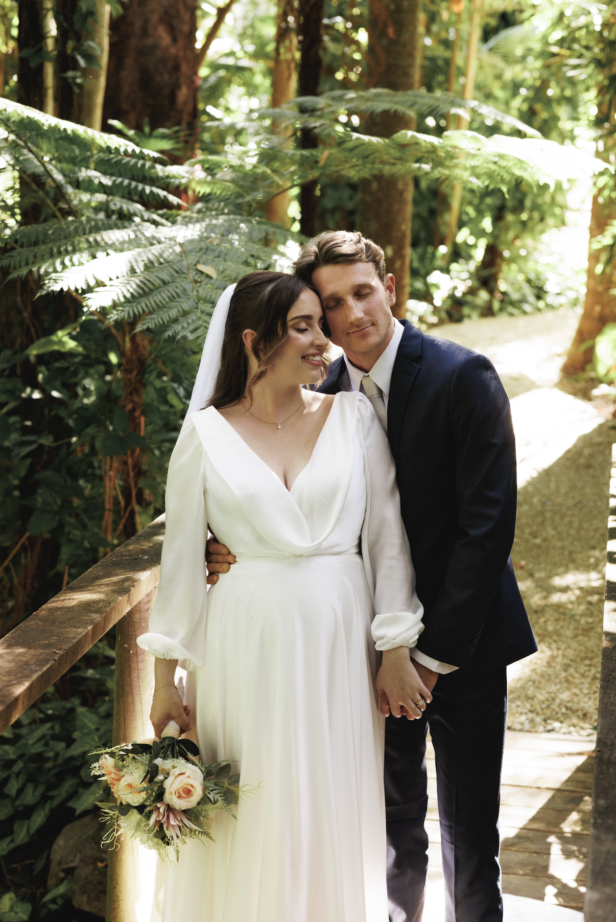 A bride and groom embracing on a forested trail, with the bride holding a bouquet of flowers and both appearing happy and serene.