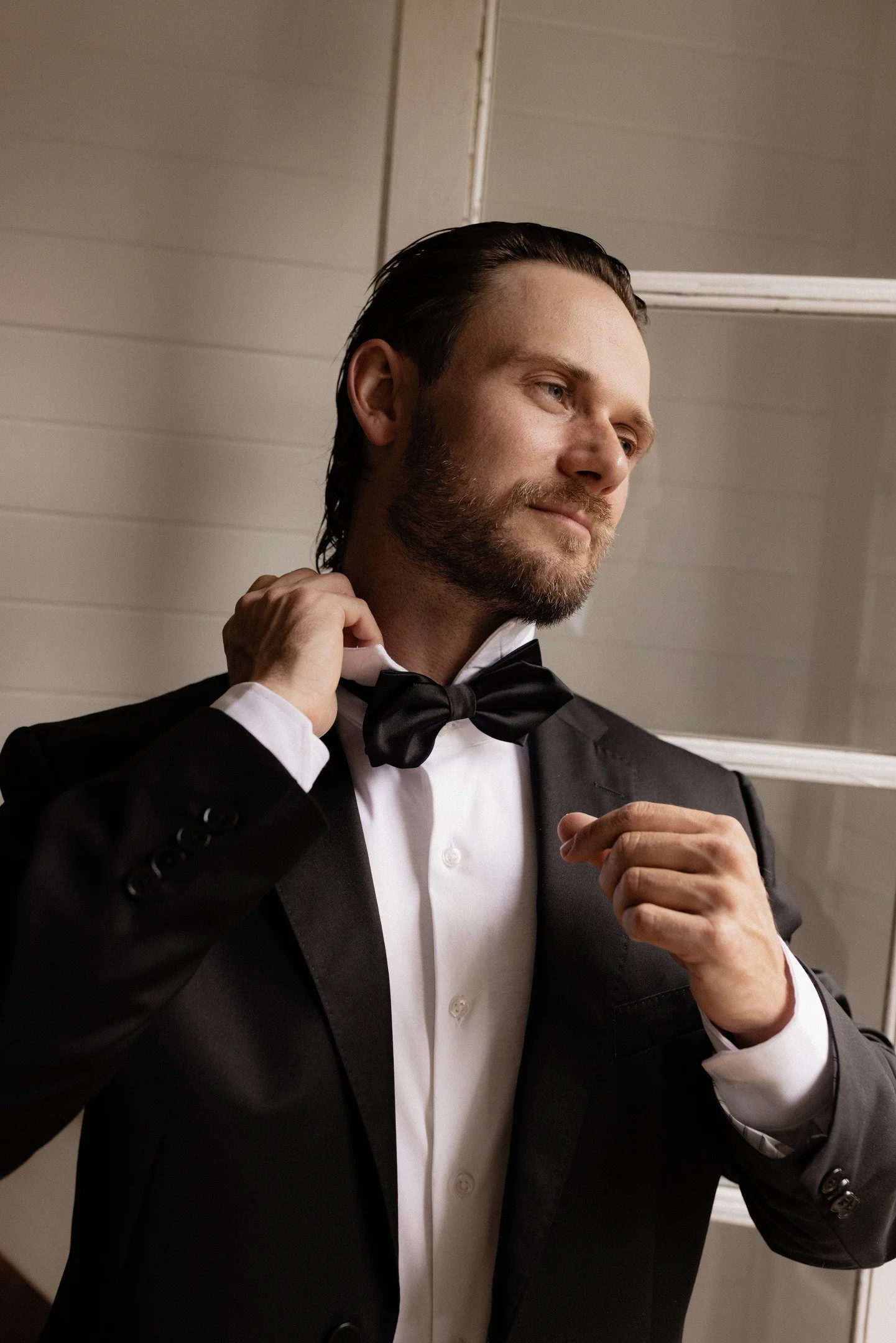 A man in a tuxedo adjusting his bowtie in front of a mirror.