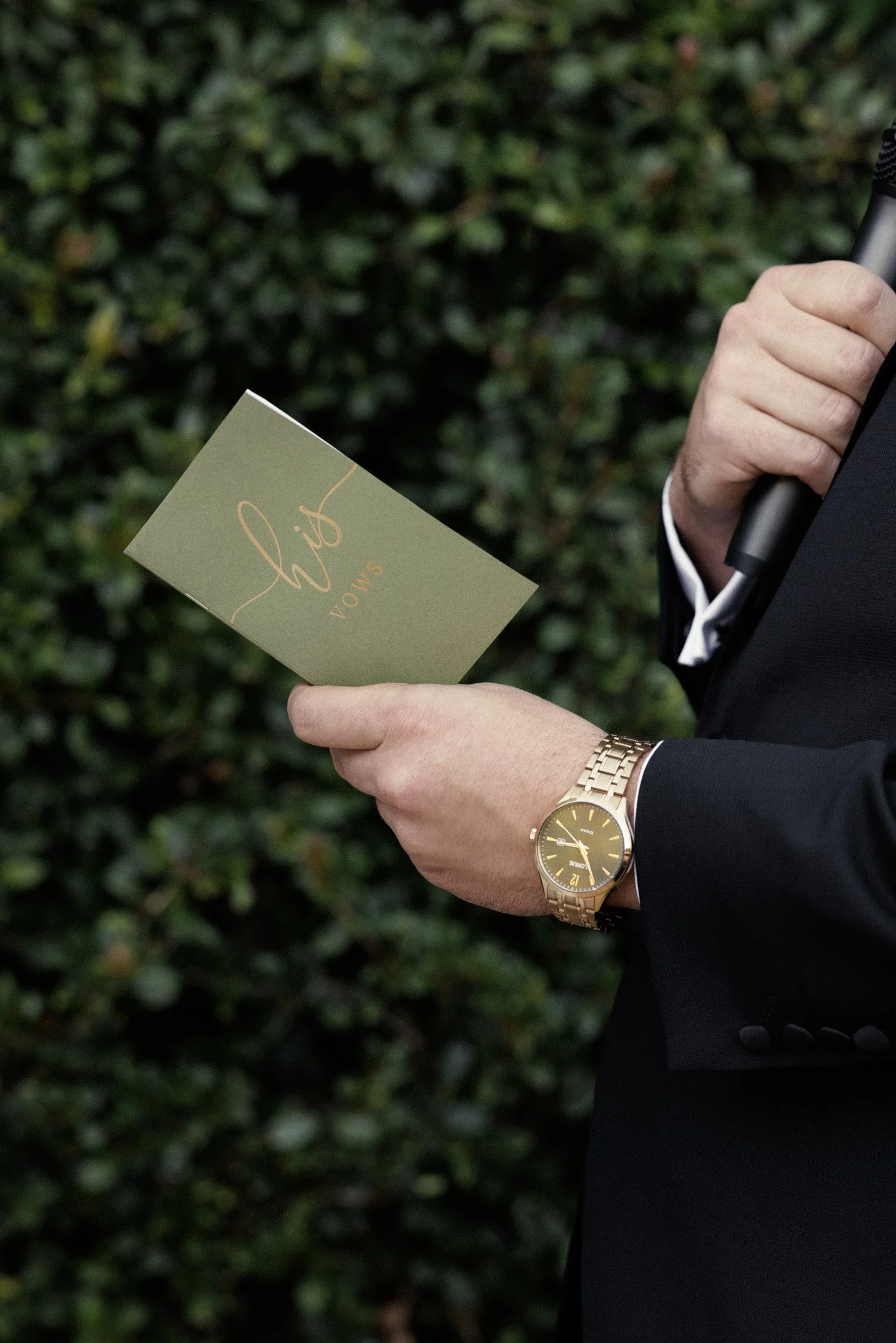 A person in a black suit holding a light green card with the brand name 'SLOANE' written on it, wearing a gold watch, standing outdoors in front of green foliage.