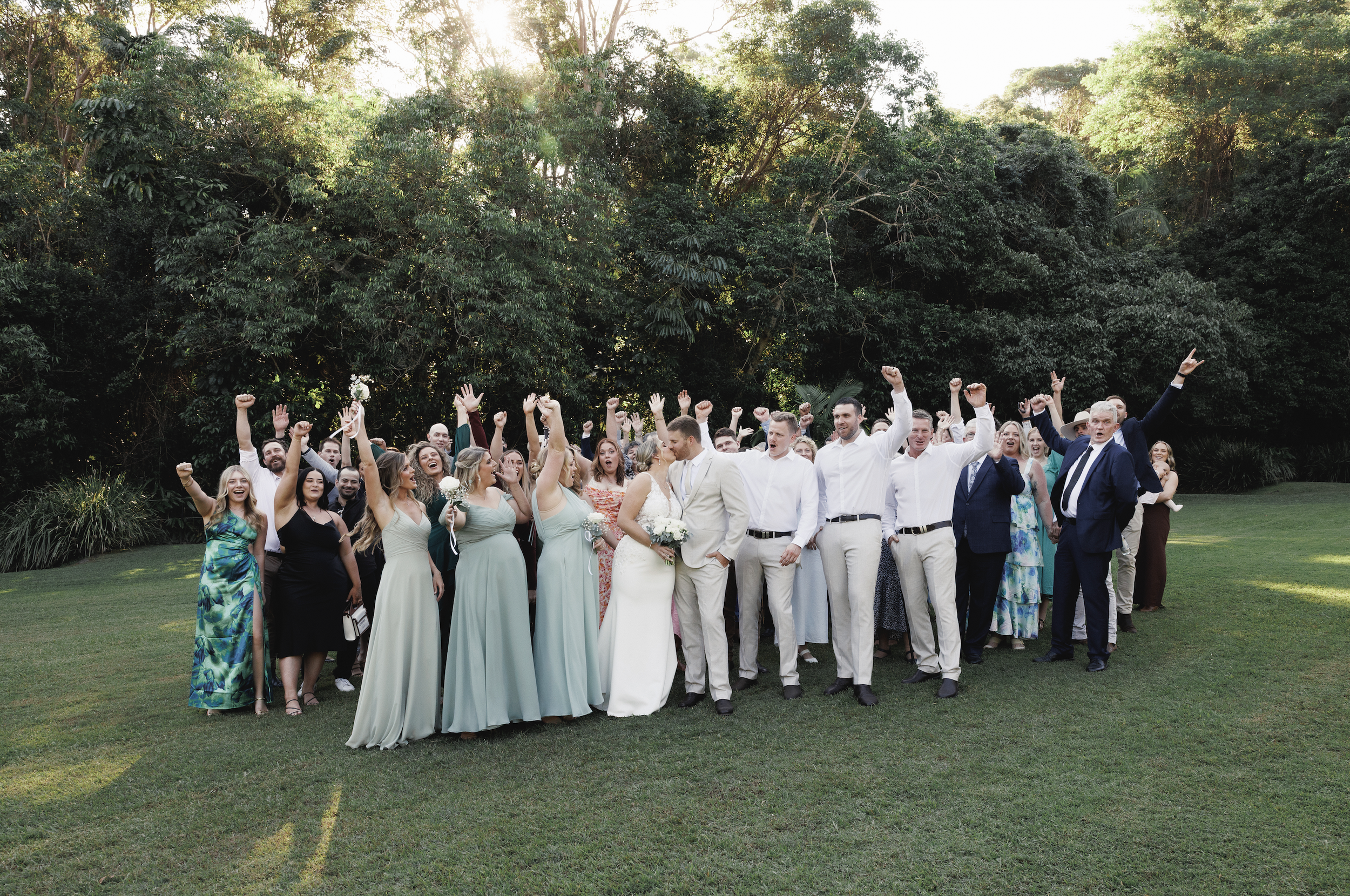 A large group of wedding guests and the bride and groom celebrate together outdoors on a grassy area with trees in the background, raising their hands in joy.