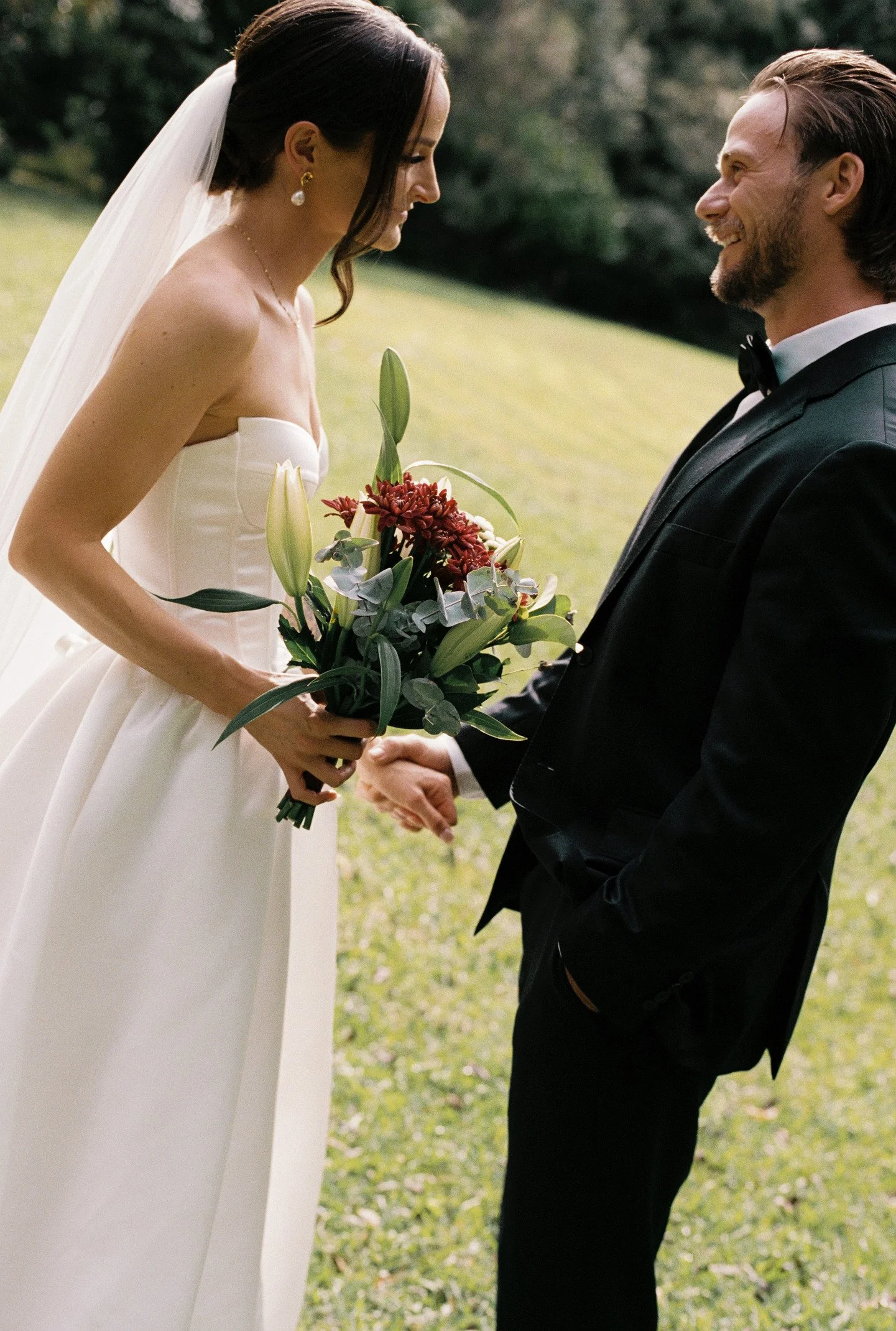 Bride in a white wedding dress holding a bouquet of flowers, standing outdoors, smiling at groom in a black tuxedo. They are holding hands, and their faces are close as they smile at each other.