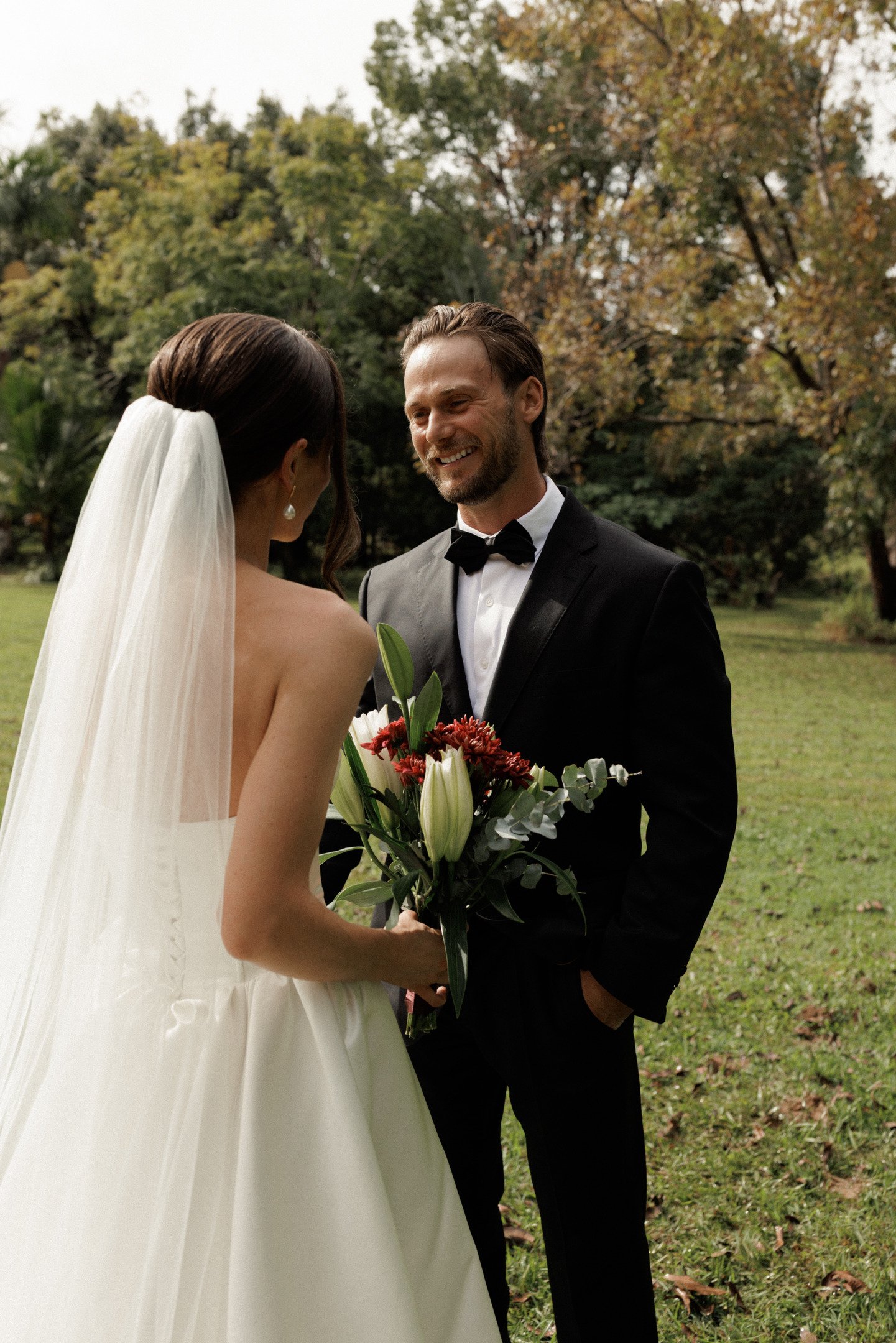 A bride and groom standing outdoors in a park, exchanging a smile at their wedding, with trees and autumn foliage in the background. The bride holds a bouquet of flowers.