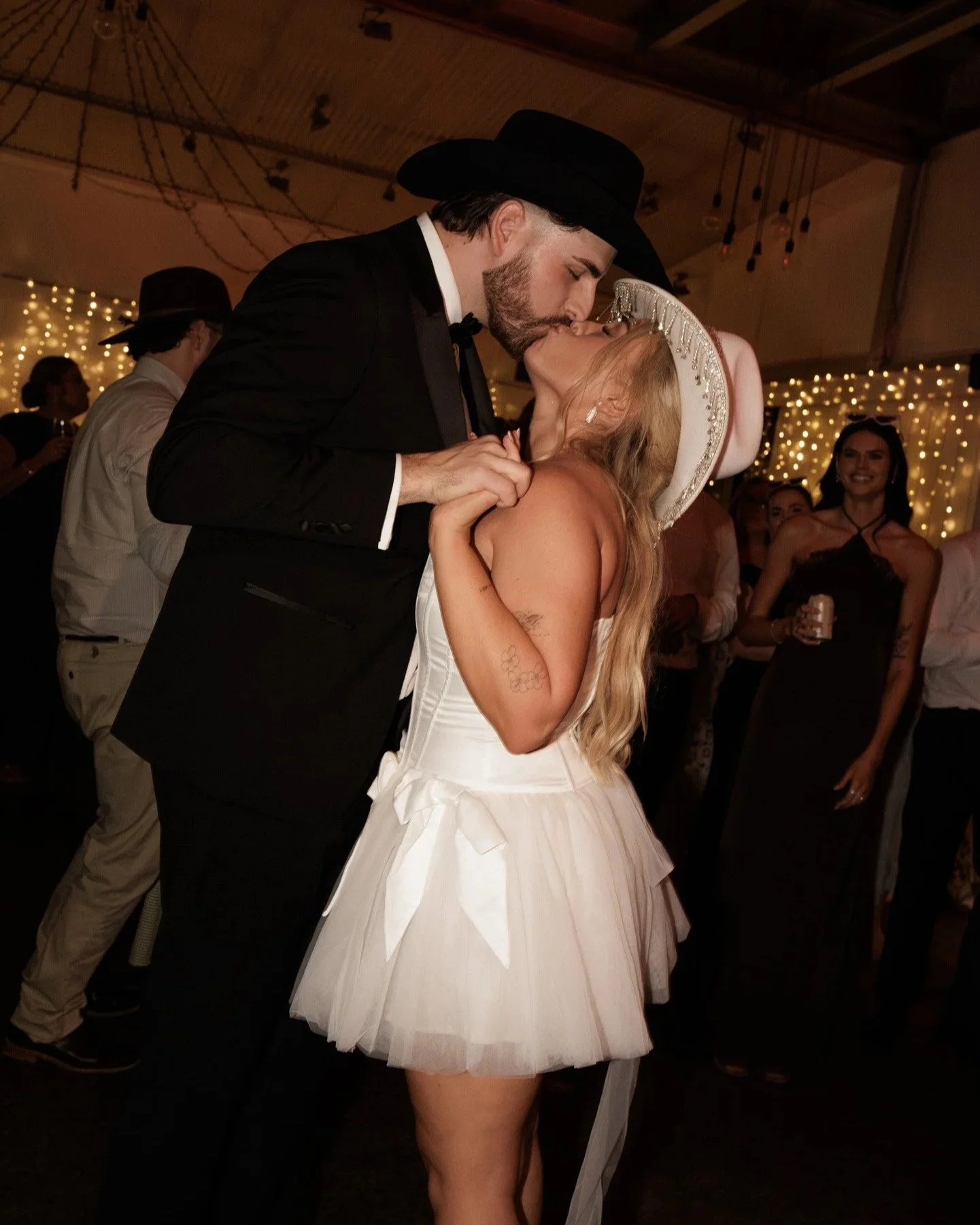 A couple kissing at a wedding reception, with the groom in a black suit and cowboy hat, and the bride in a white dress and matching wide-brimmed hat. Guests and decorative lights are visible in the background.