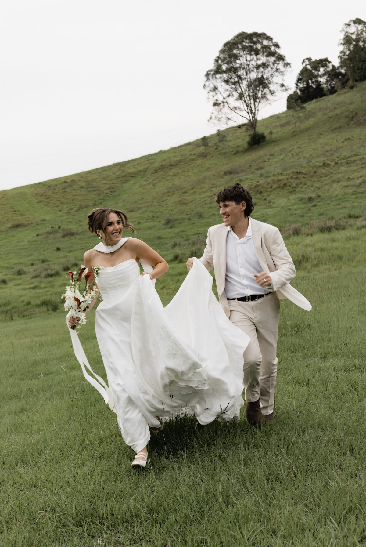 A newlywed couple running and smiling across a grassy field with green hills in the background, one holding a bouquet, in overcast weather.