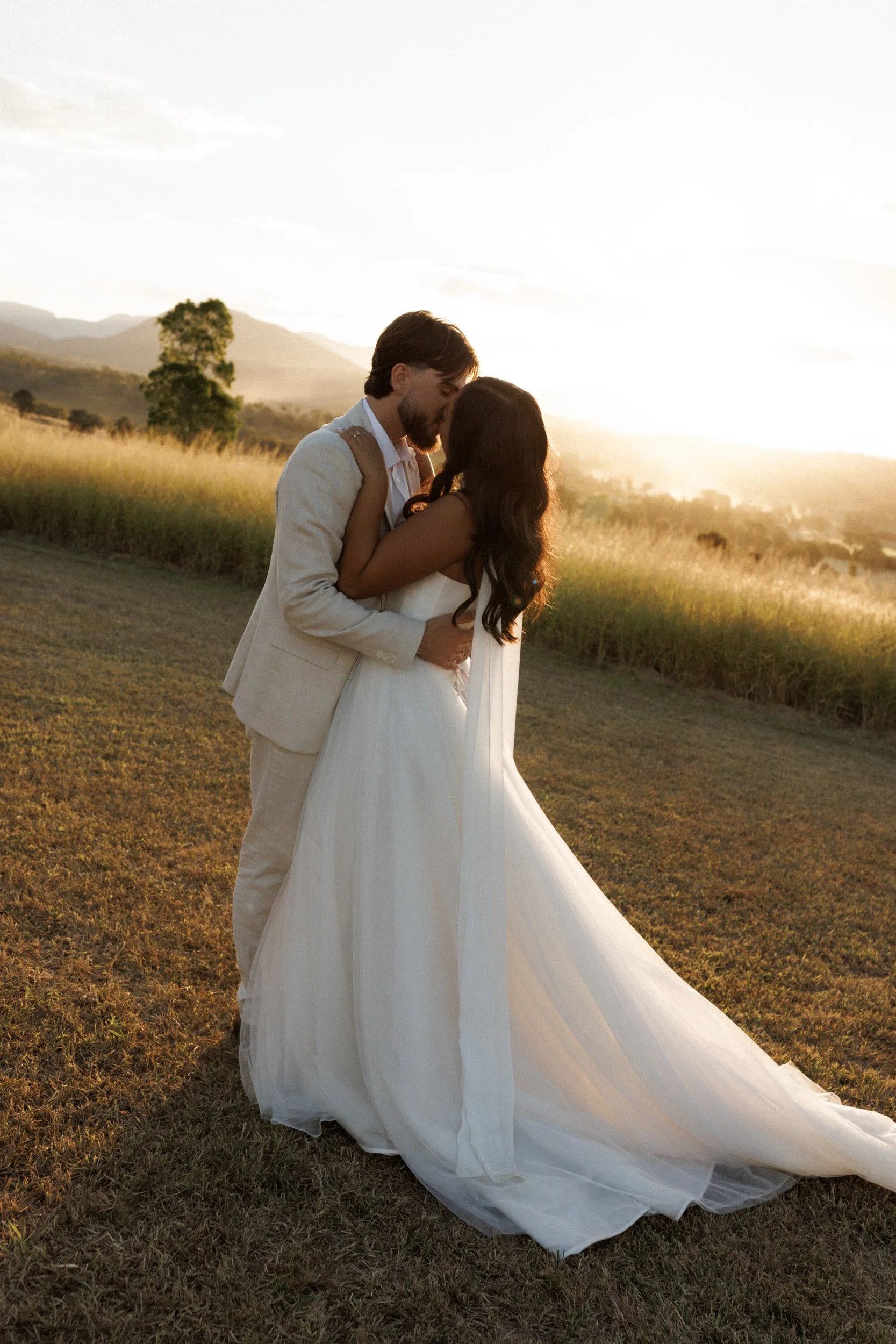 A couple dressed in wedding attire sharing a kiss outdoors at sunset, with wide-open field, mountains, and a single tree in the background.