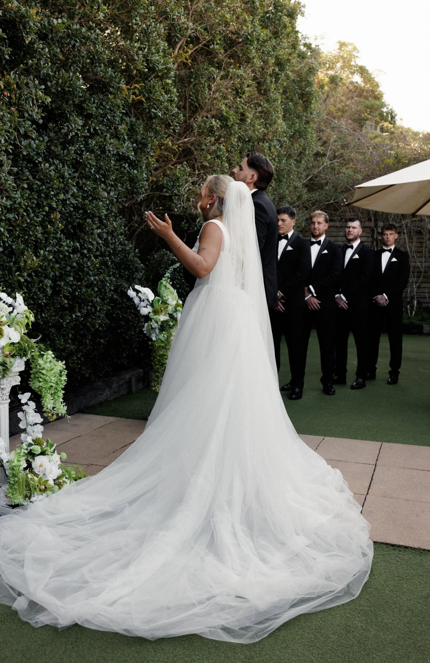 A bride and groom at a wedding ceremony outdoors, with groomsmen in tuxedos standing in the background, and a lush hedge and floral decorations in the setting.