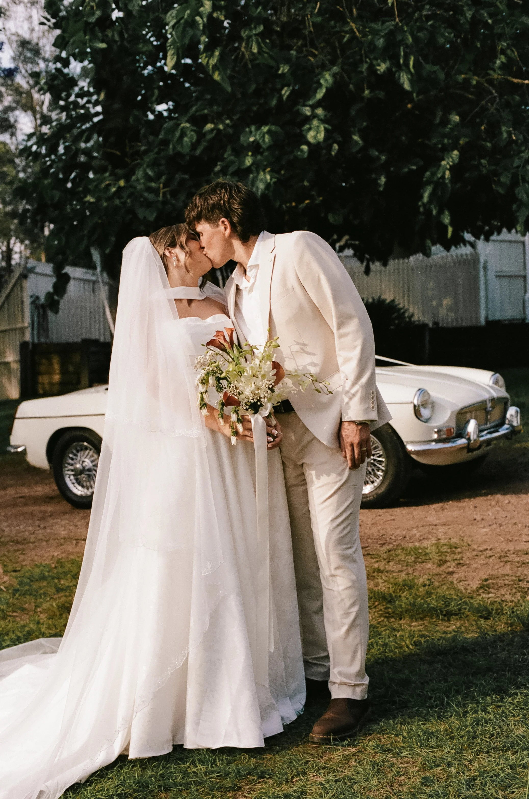 A bride and groom sharing a kiss outdoors next to a vintage white car, with a large tree in the background.