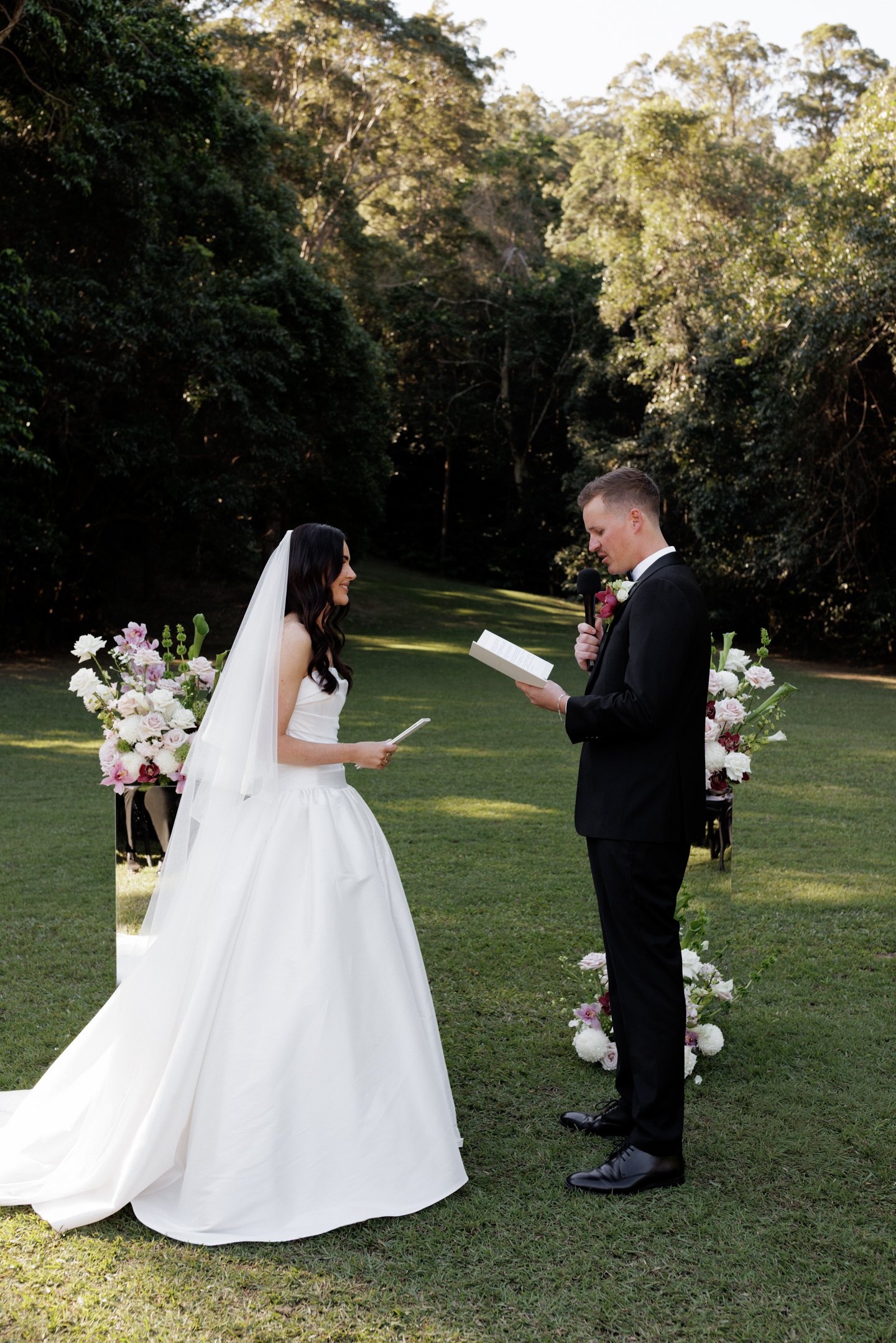 A bride and groom stand in a garden during their wedding ceremony. The bride wears a white wedding gown and veil, while the groom wears a black tuxedo with a boutonniere. The groom holds a microphone and a book, reading vows or a speech. Floral arran