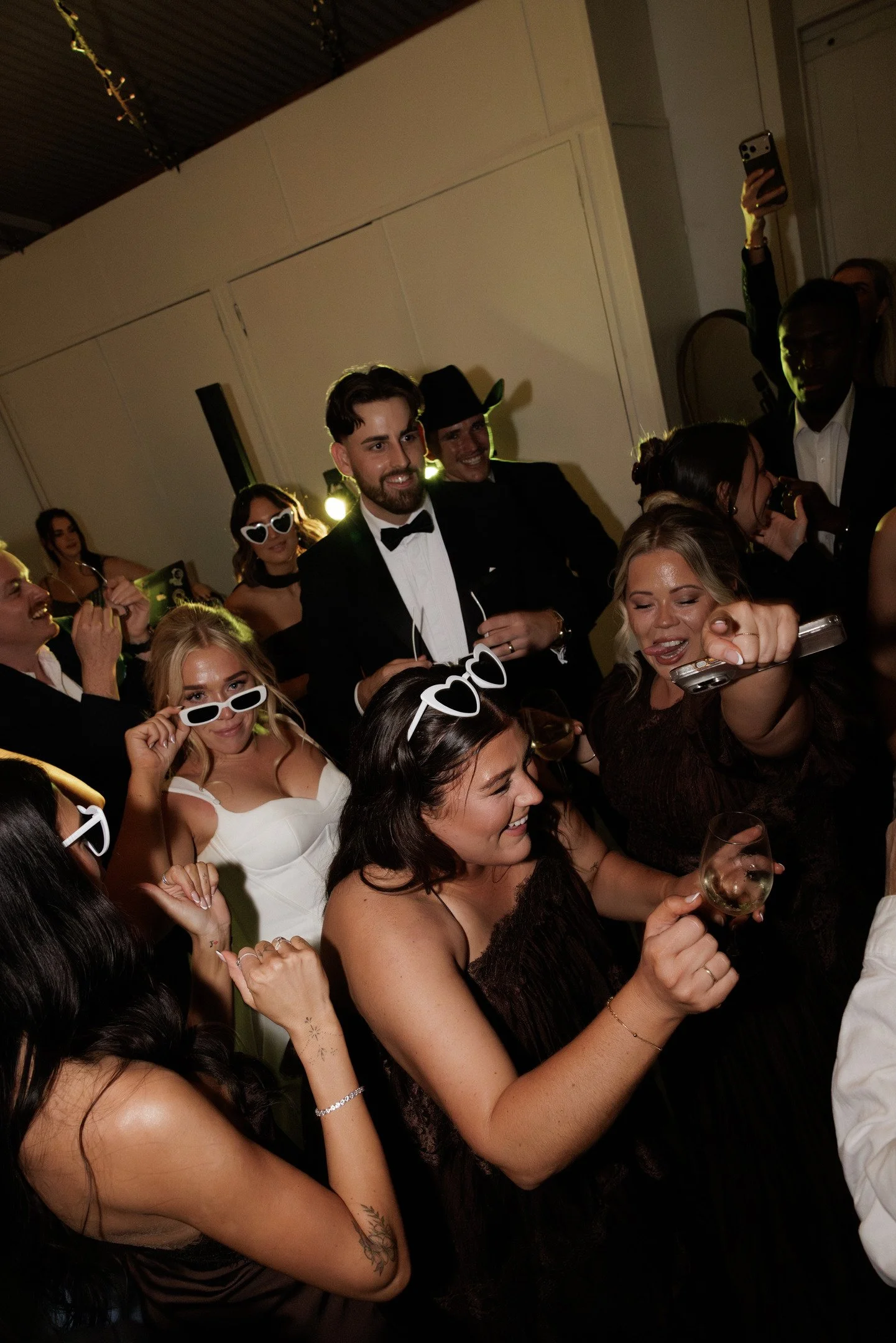 People at a party, some wearing sunglasses and formal attire, holding drinks, dancing, and taking selfies.