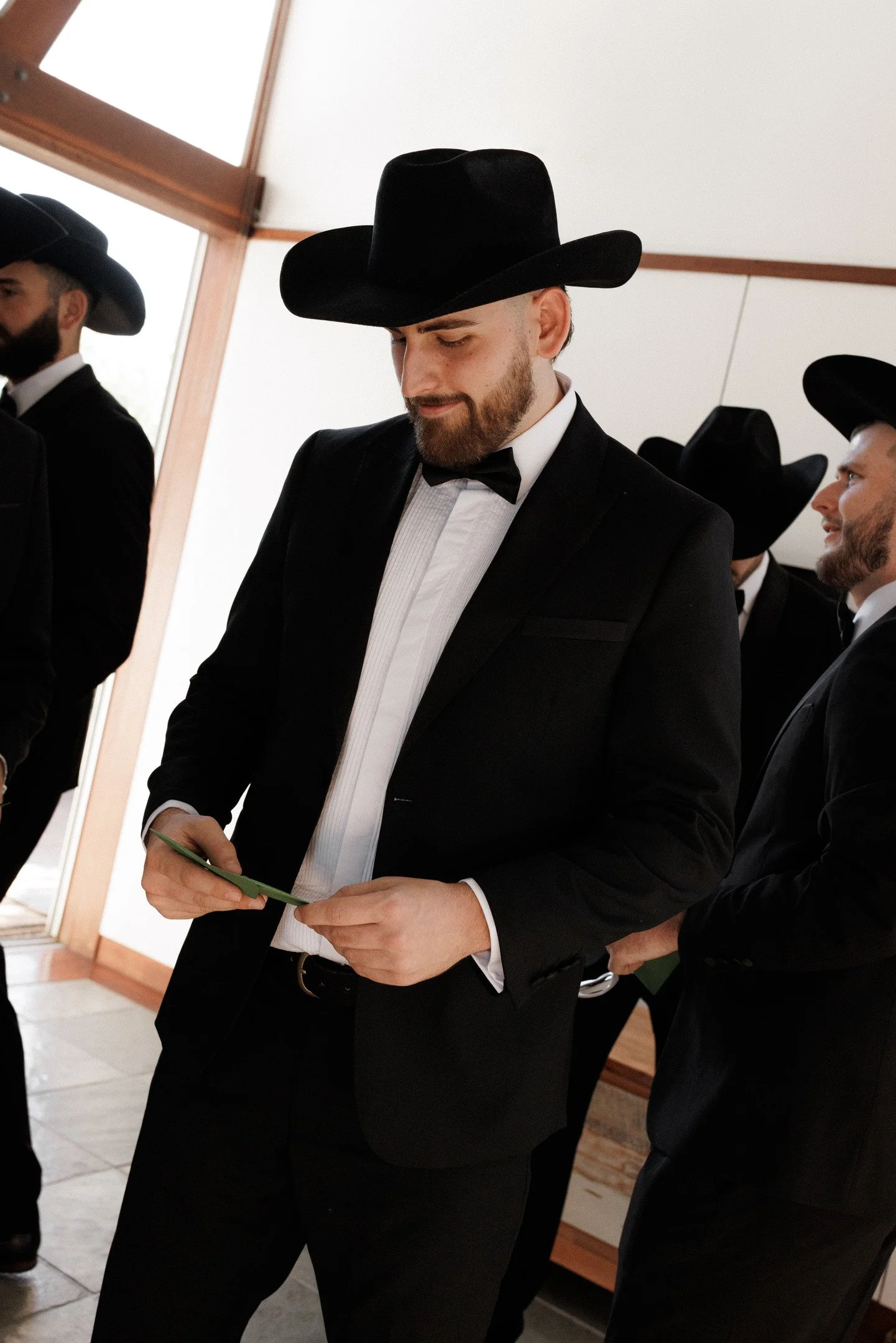 A group of men dressed in black tuxedos and wearing large black cowboy hats, standing indoors.