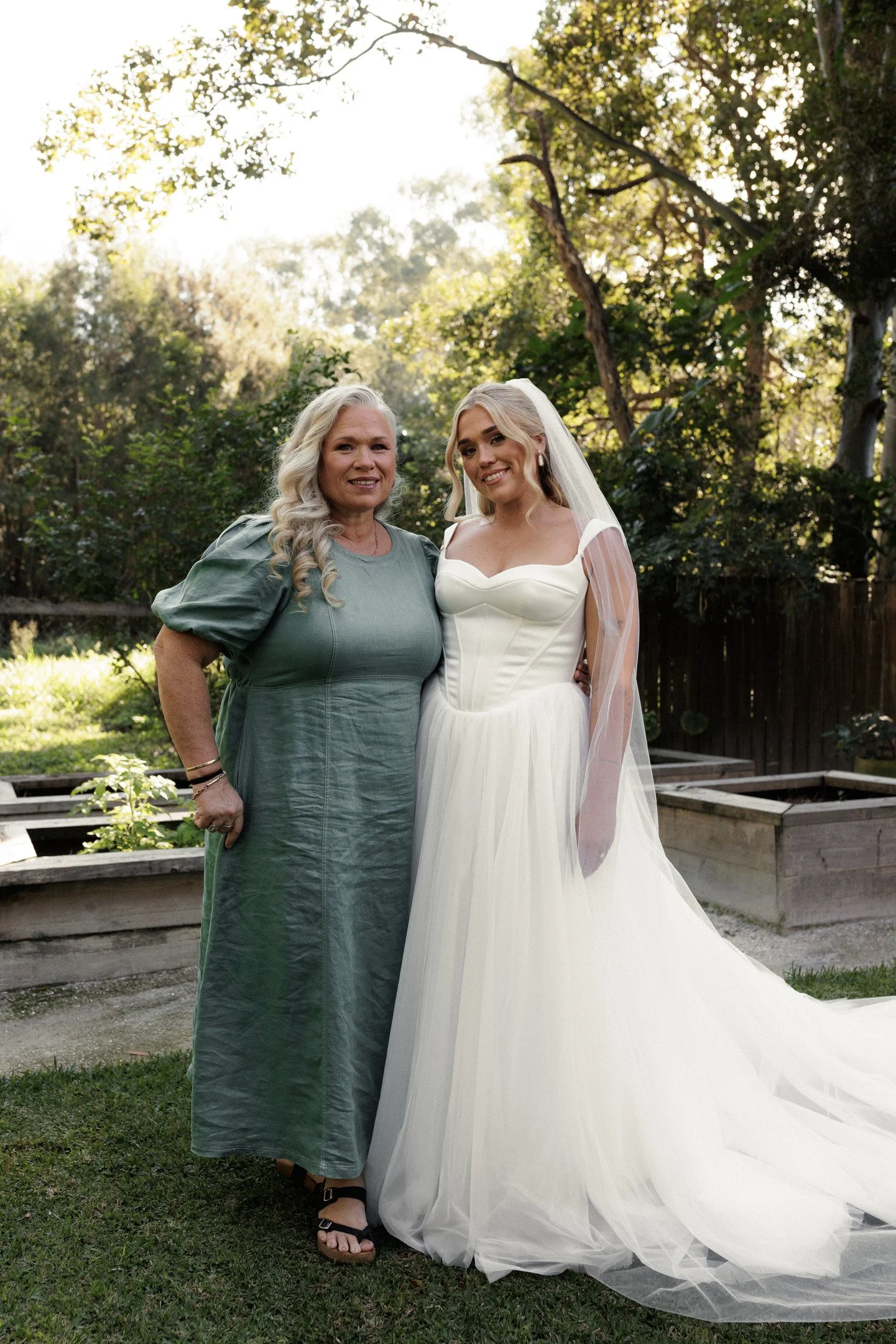 Two women standing outdoors in a garden, one in a light green dress with long blonde curly hair, and the other in a white wedding dress with a veil, smiling at the camera, surrounded by trees and sunlight filtering through the leaves.