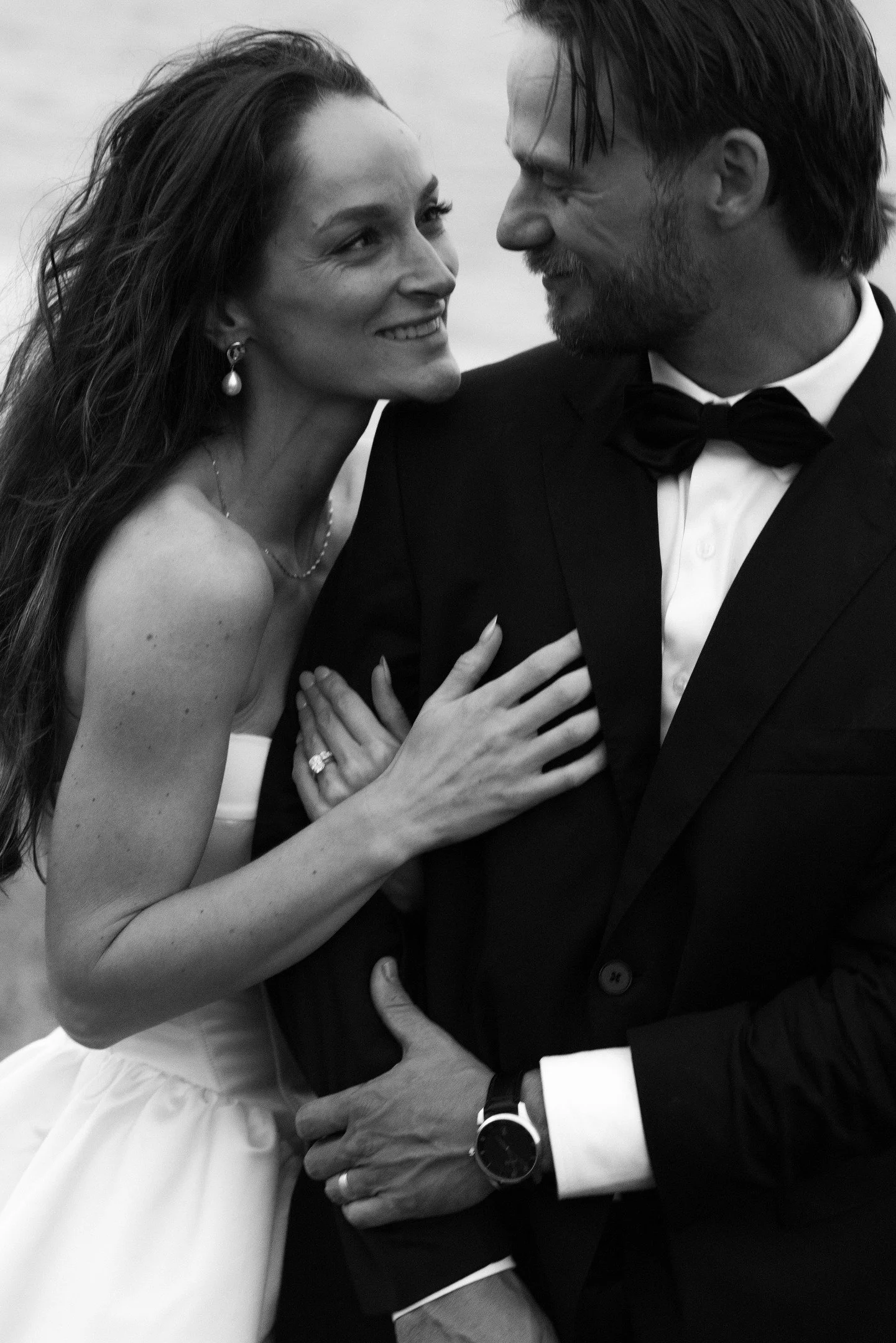 A black and white photo of a man and woman dressed in formal attire, smiling closely and looking into each other's eyes, with hands touching each other.