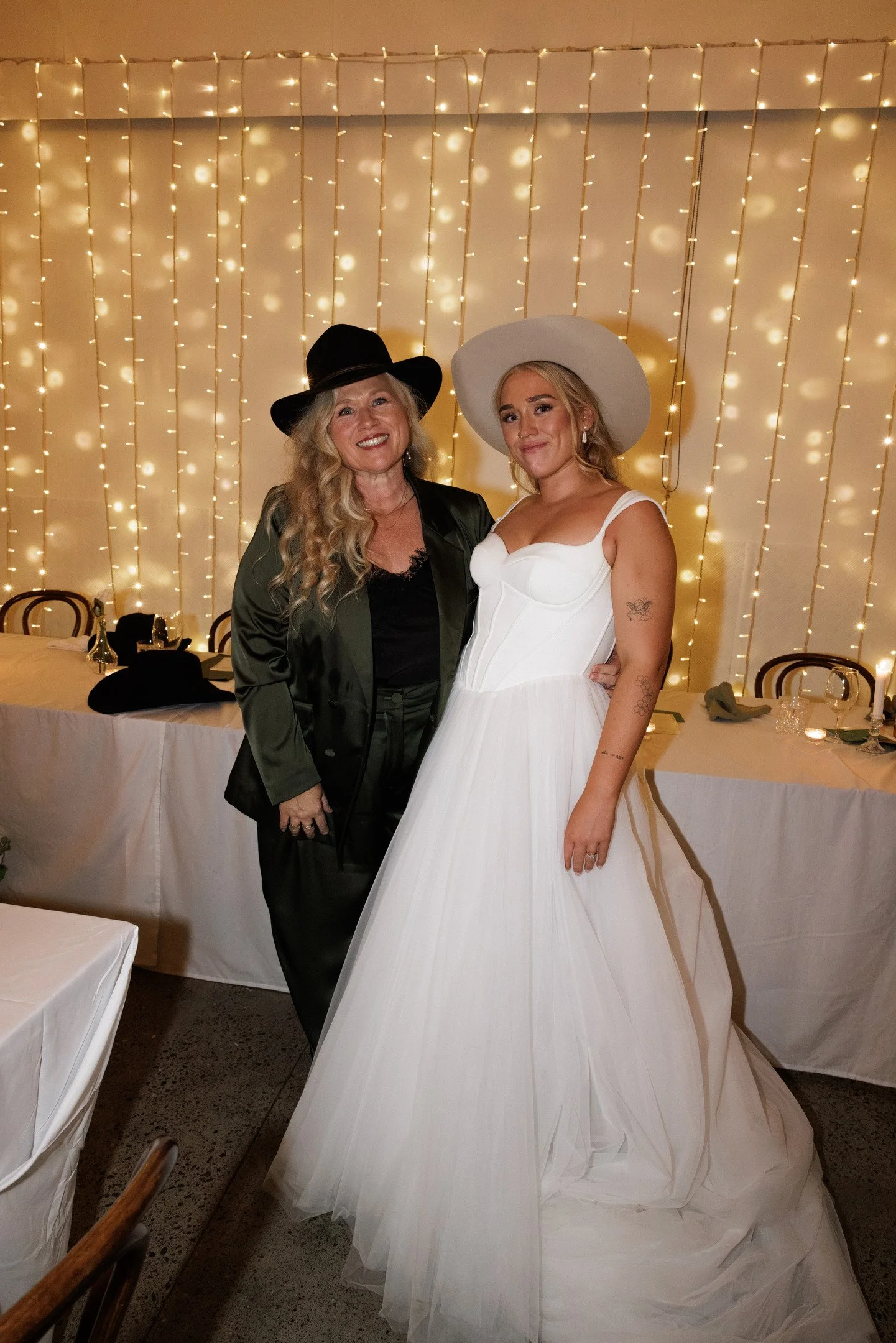 Two women, one older and one younger, standing together at an event with string lights in the background. The older woman is wearing a black hat and a dark blazer, while the younger woman is in a white dress and wide-brimmed hat. They are smiling and