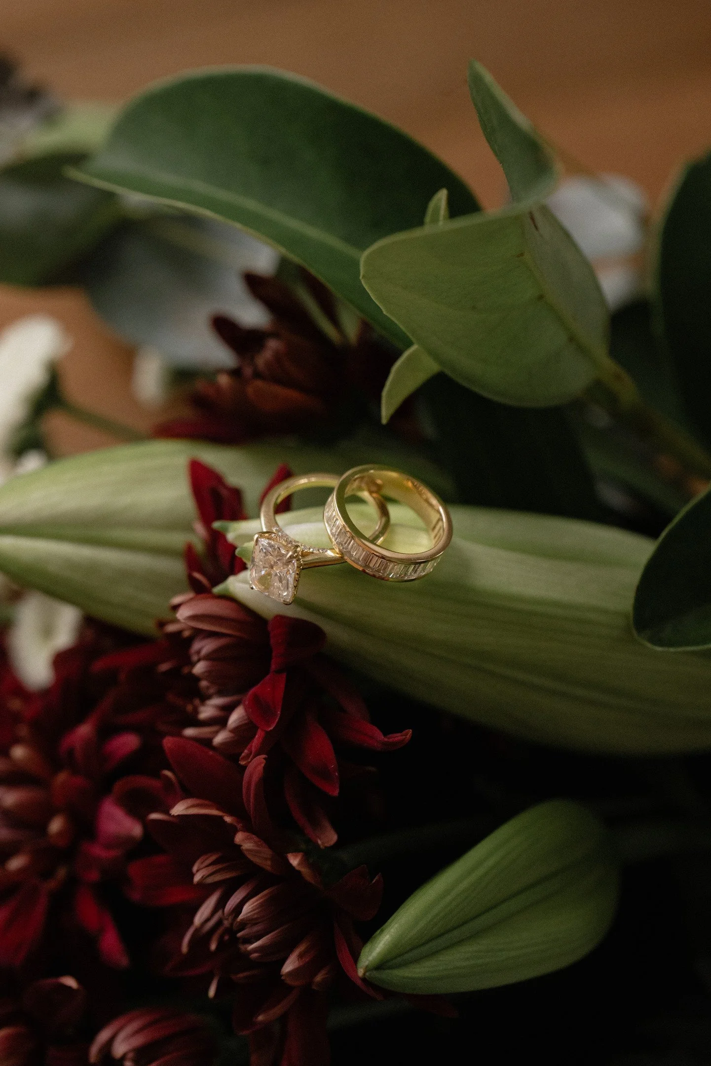 A close-up photograph of a wedding ring and engagement ring resting on a bouquet of flowers and green leaves.