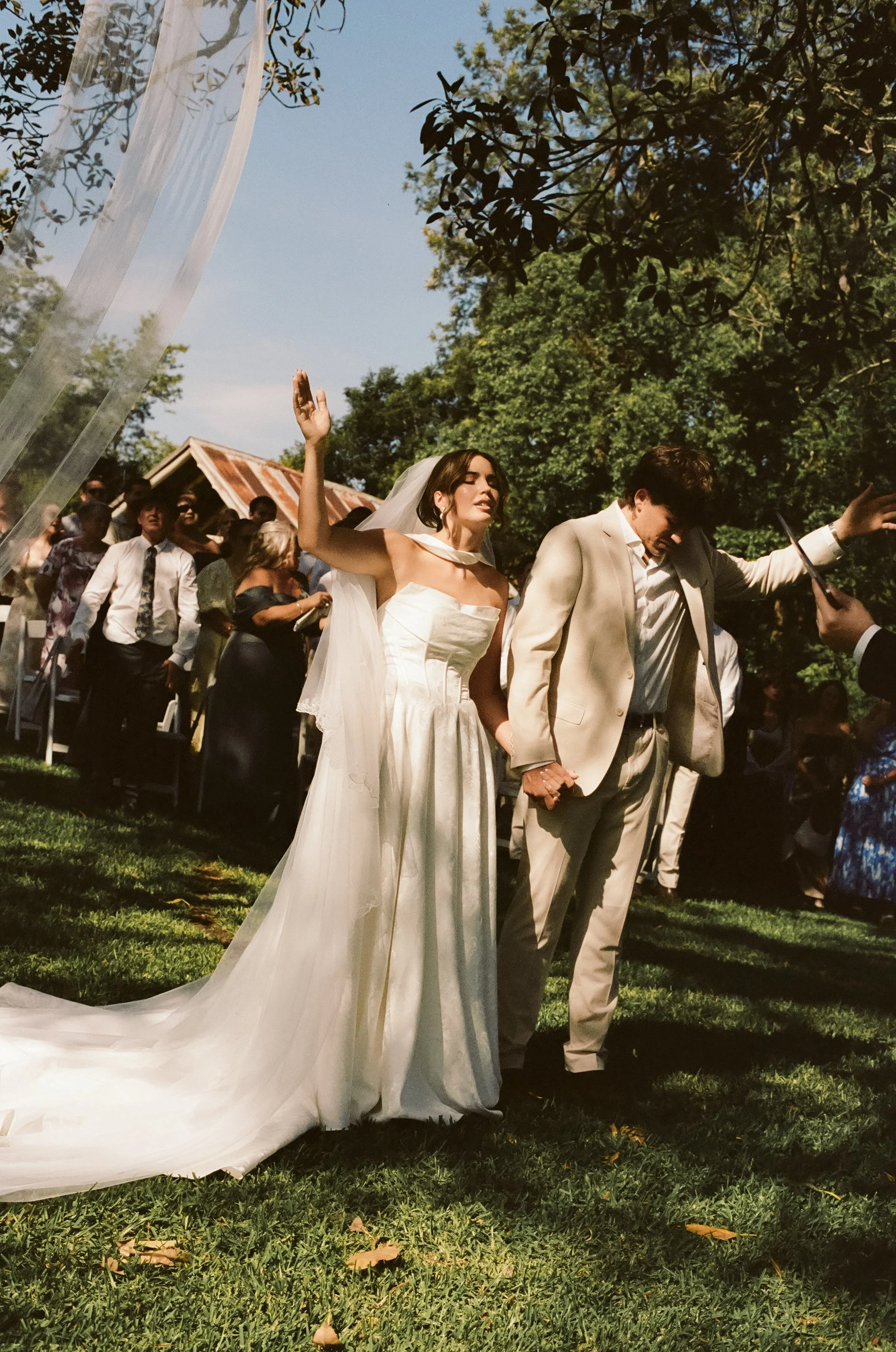 Bride and groom holding hands during an outdoor wedding ceremony with guests seated in the background.