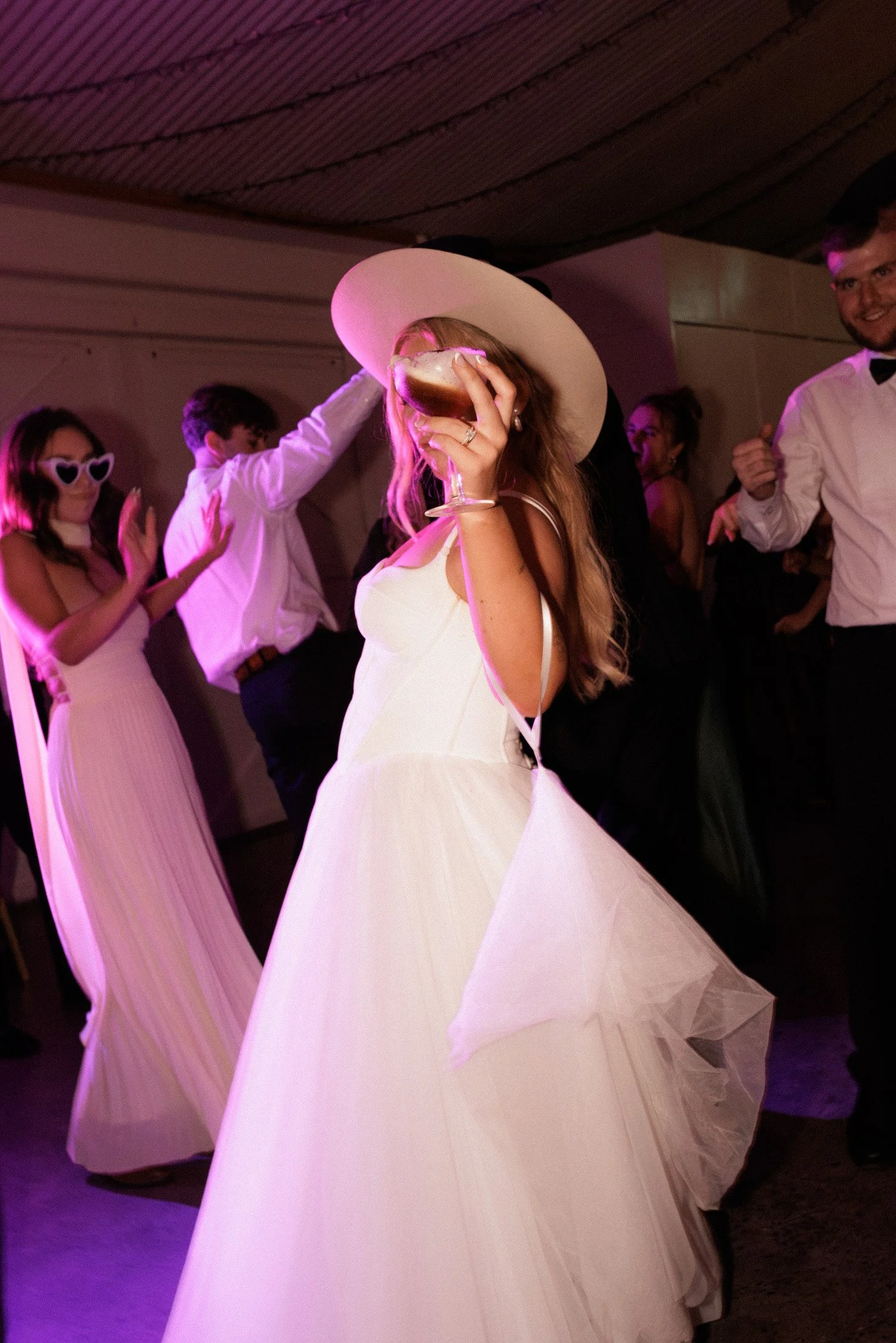 A woman in a white dress and wide-brimmed hat dancing at a party with other guests, some wearing sunglasses and formal attire.