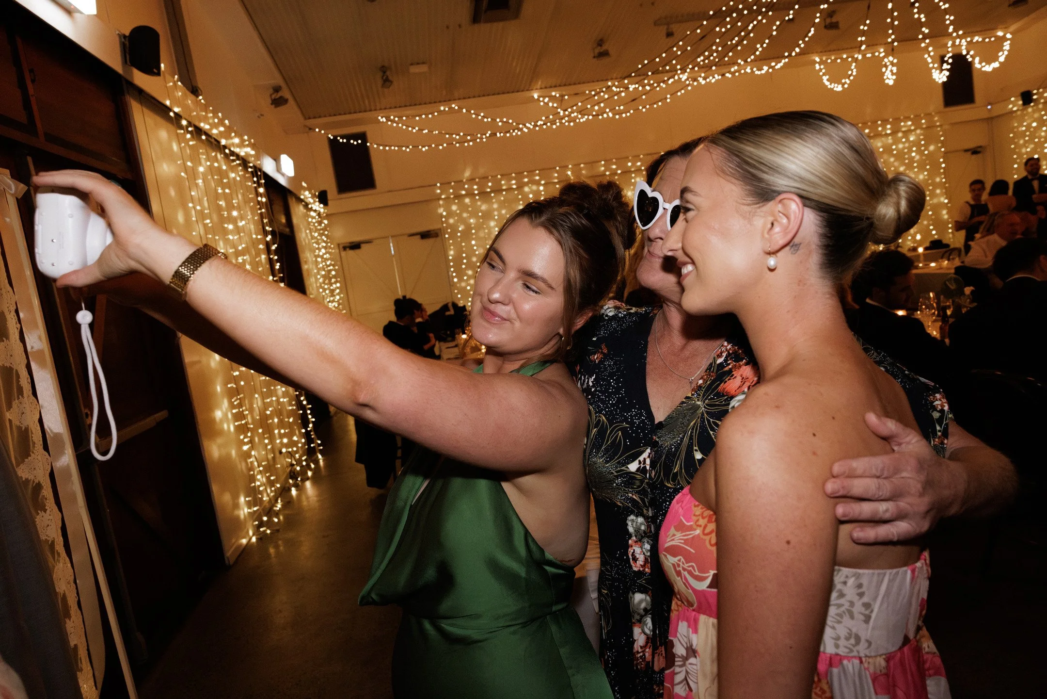 Three women at a celebration are taking a selfie together, with one woman holding a camera. They are behind a wall decorated with string lights. The women are smiling and dressed in festive clothing, and the background shows other guests and a decora