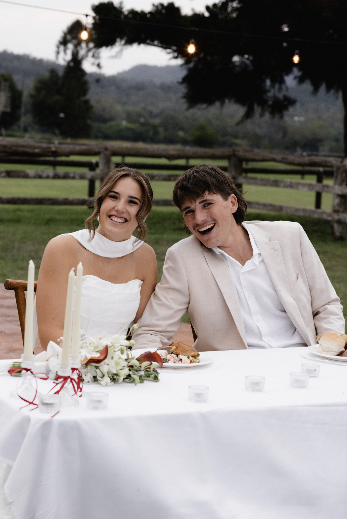 A smiling bride and groom sitting at a table outdoors during their wedding reception, with trees and hills in the background and string lights overhead.