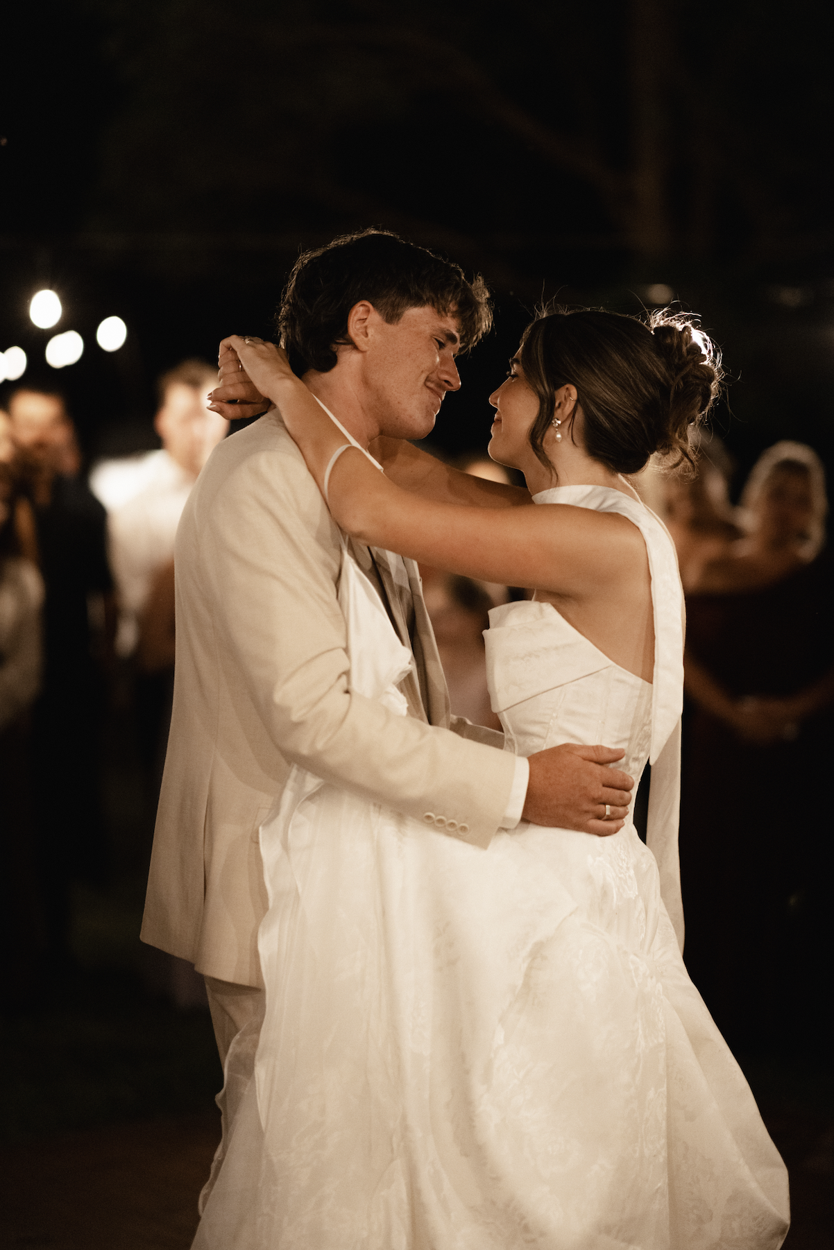 A couple, dressed in wedding attire, dancing closely during their wedding reception at night, with blurred guests in the background.