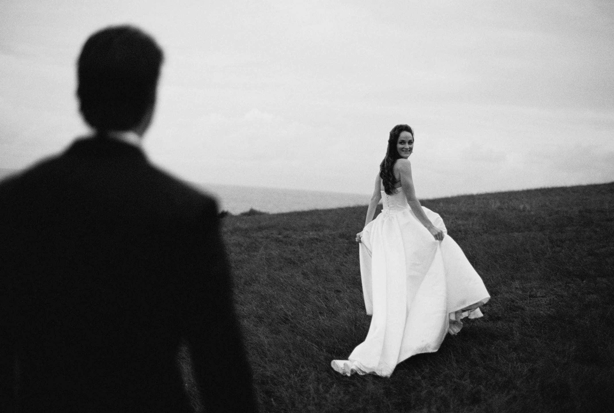 A black and white photo of a woman in a wedding dress smiling and sitting on the grass outdoors, with a blurred man in a suit in the foreground.