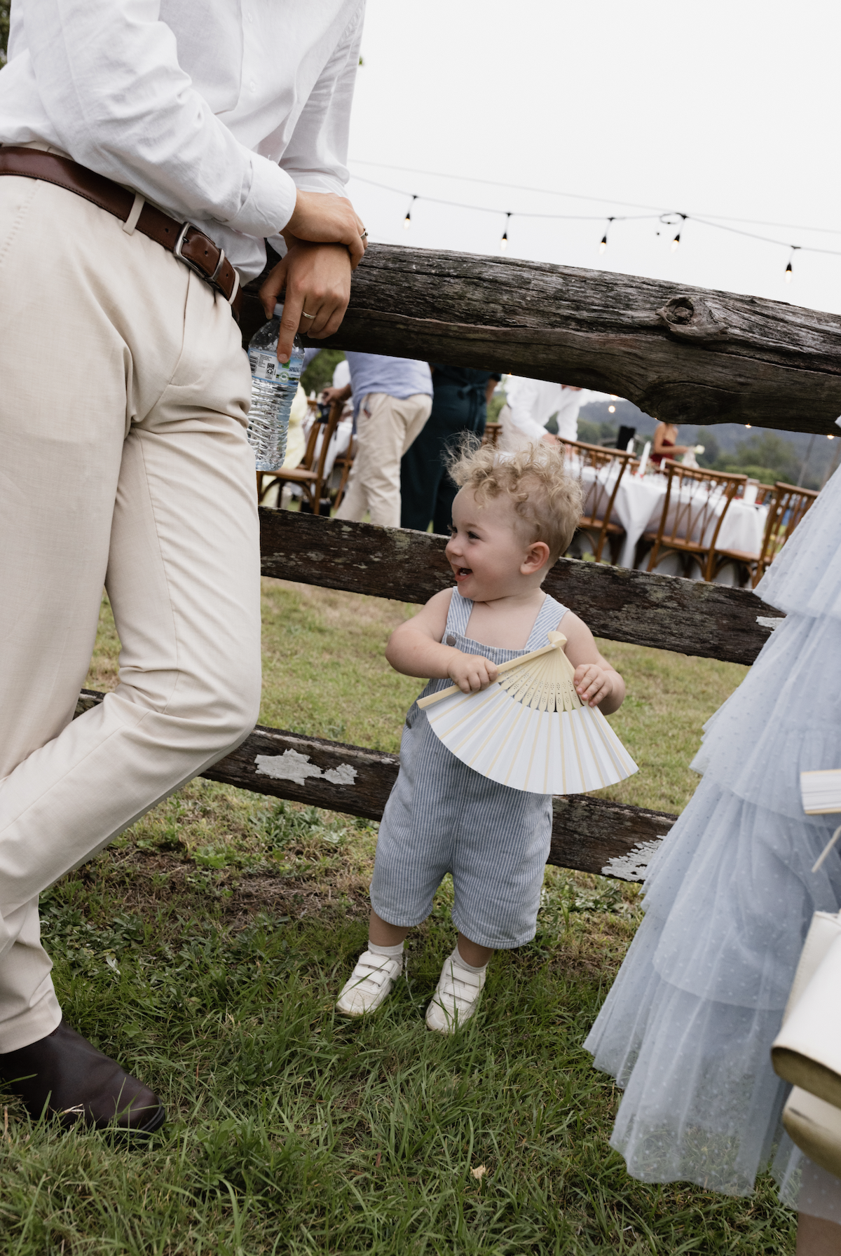 A young child with curly blonde hair, wearing light blue striped overalls and white shoes, holding a handheld fan, and smiling as they look at an adult in white pants and a white shirt at an outdoor event with tables and string lights in the backgrou