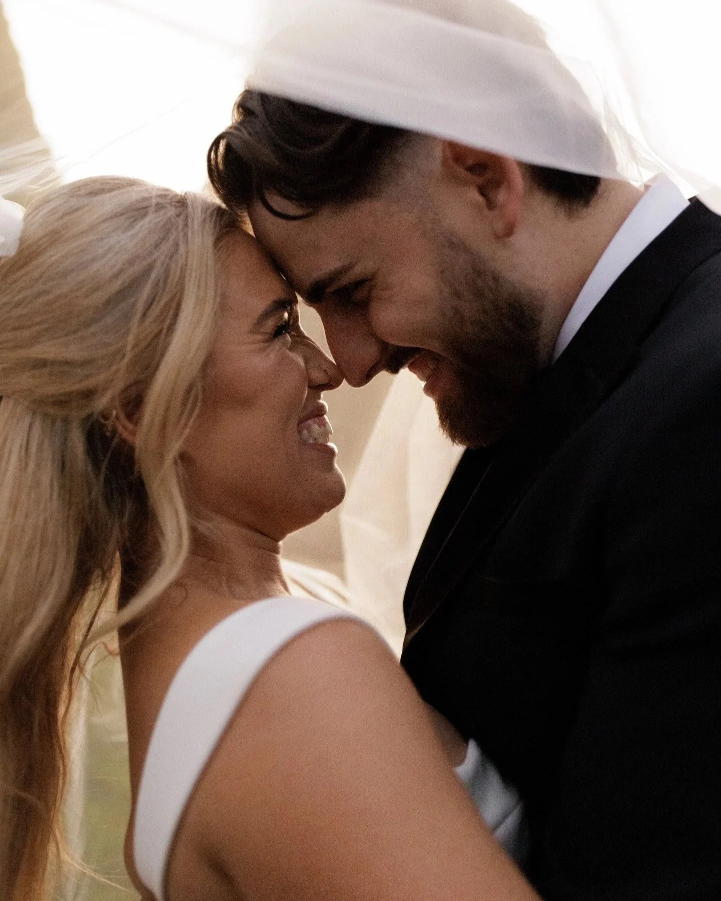 A close-up of a happy couple touching foreheads, smiling at each other, with the woman having long blonde hair and the man wearing a white chef's hat and black suit, in a warm, intimate moment.