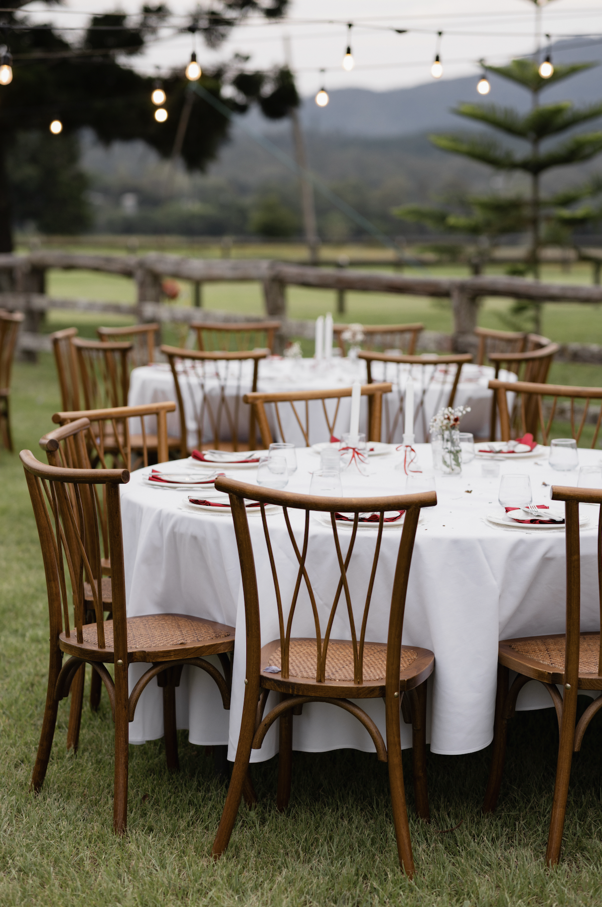 Outdoor wedding reception setup with round tables covered with white tablecloths, decorated with candles and small flower arrangements, surrounded by wooden chairs, string lights overhead, and a rustic fence in the background, with mountains and tree
