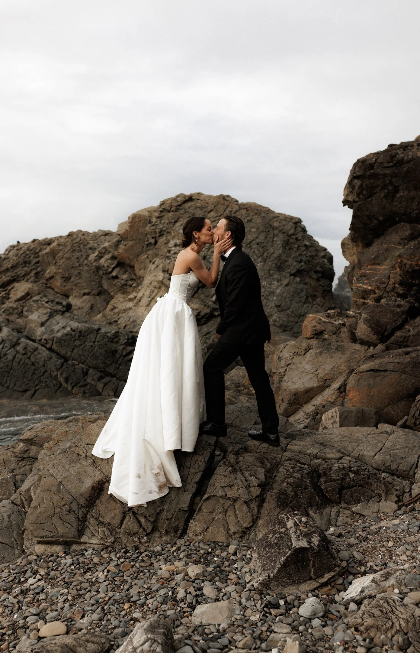 A bride and groom kiss on rocky beach, surrounded by large rocks and pebbles, overcast sky.