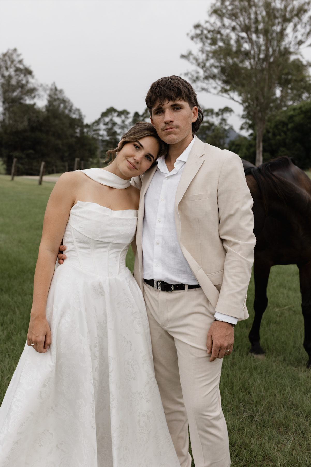 A bride and groom standing closely together outdoors on a cloudy day, with a horse partially visible behind them; the bride is wearing a white wedding dress and the groom a beige suit.