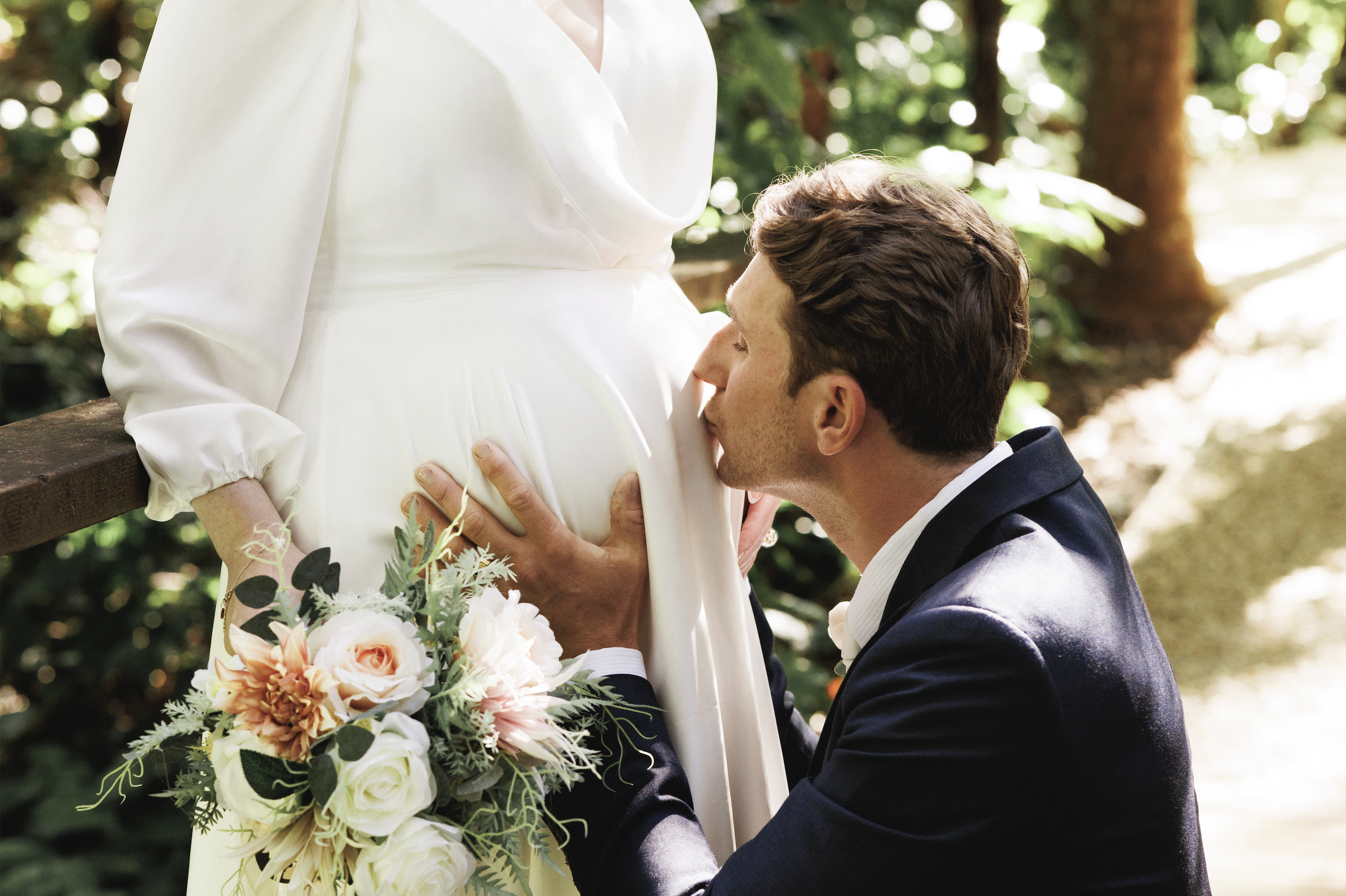 A man in a black suit and a woman in a white dress are outdoors, with the man kissing the woman's pregnant belly. The woman is holding a bouquet of flowers and standing by a wooden railing, surrounded by lush greenery.