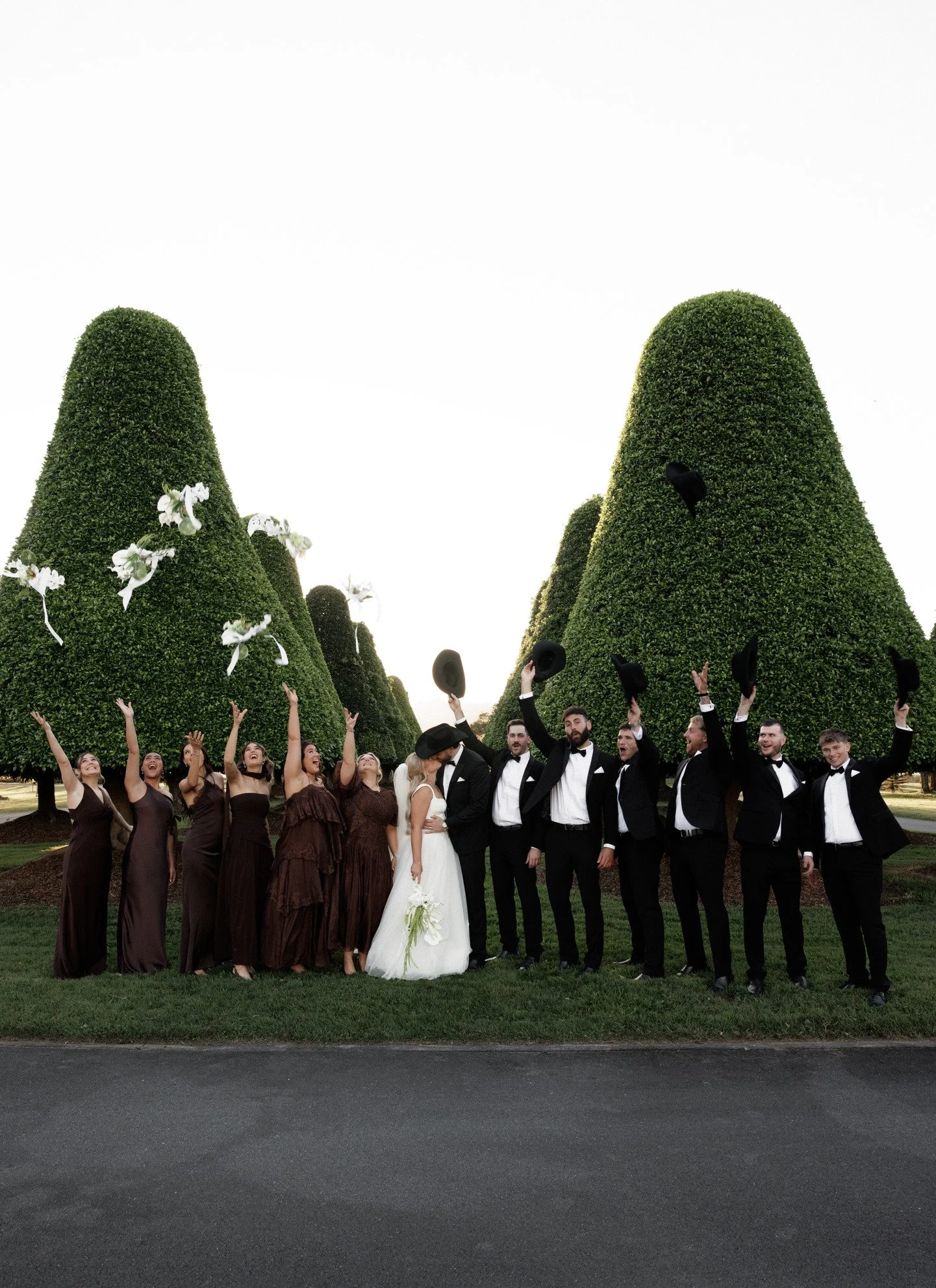 A group of people dressed in formal attire at a wedding, standing in front of neatly trimmed green bushes, some holding black hats, celebrating and smiling.