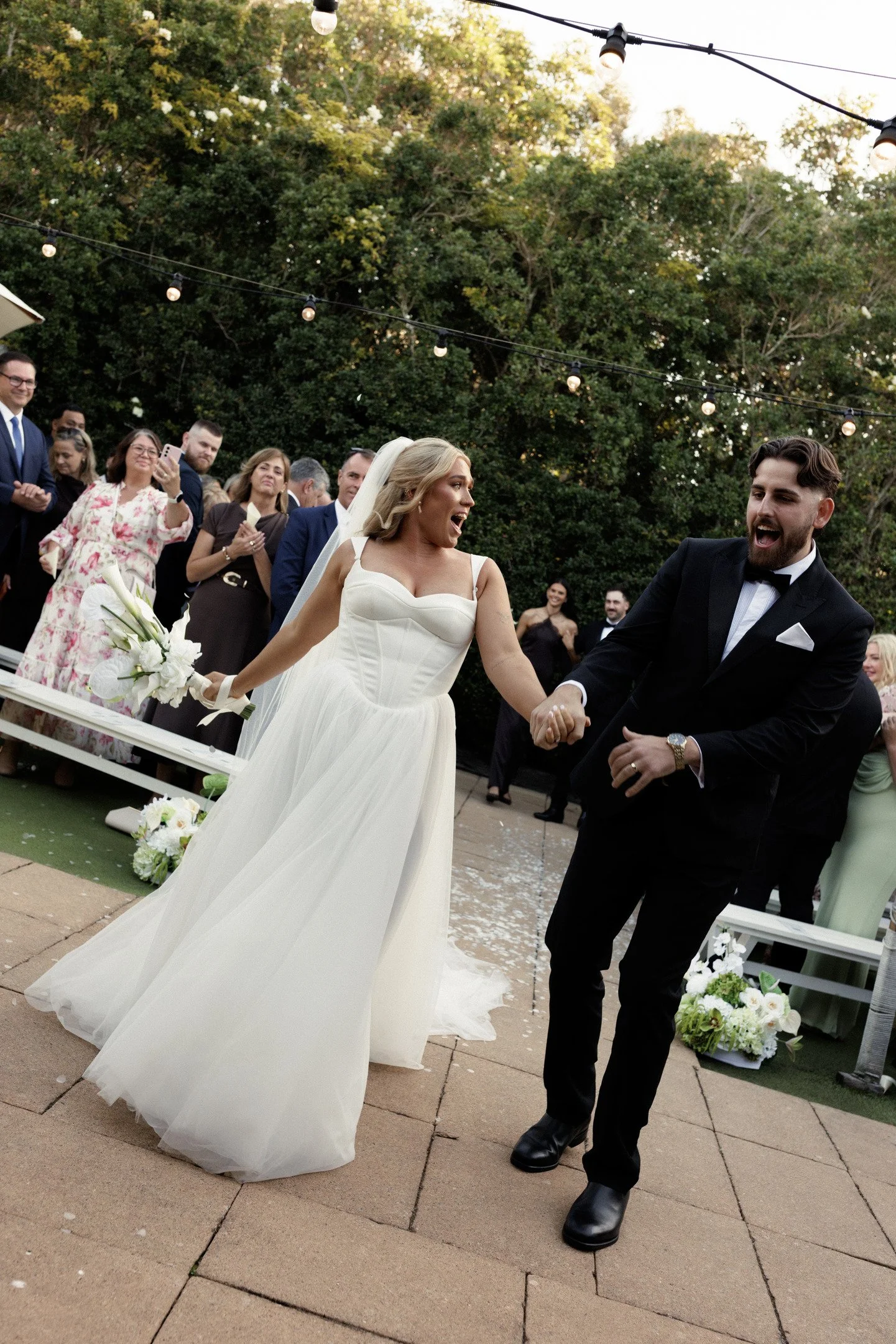 A bride and groom dancing at an outdoor wedding reception, with guests in the background, string lights overhead, and trees surrounding the area.