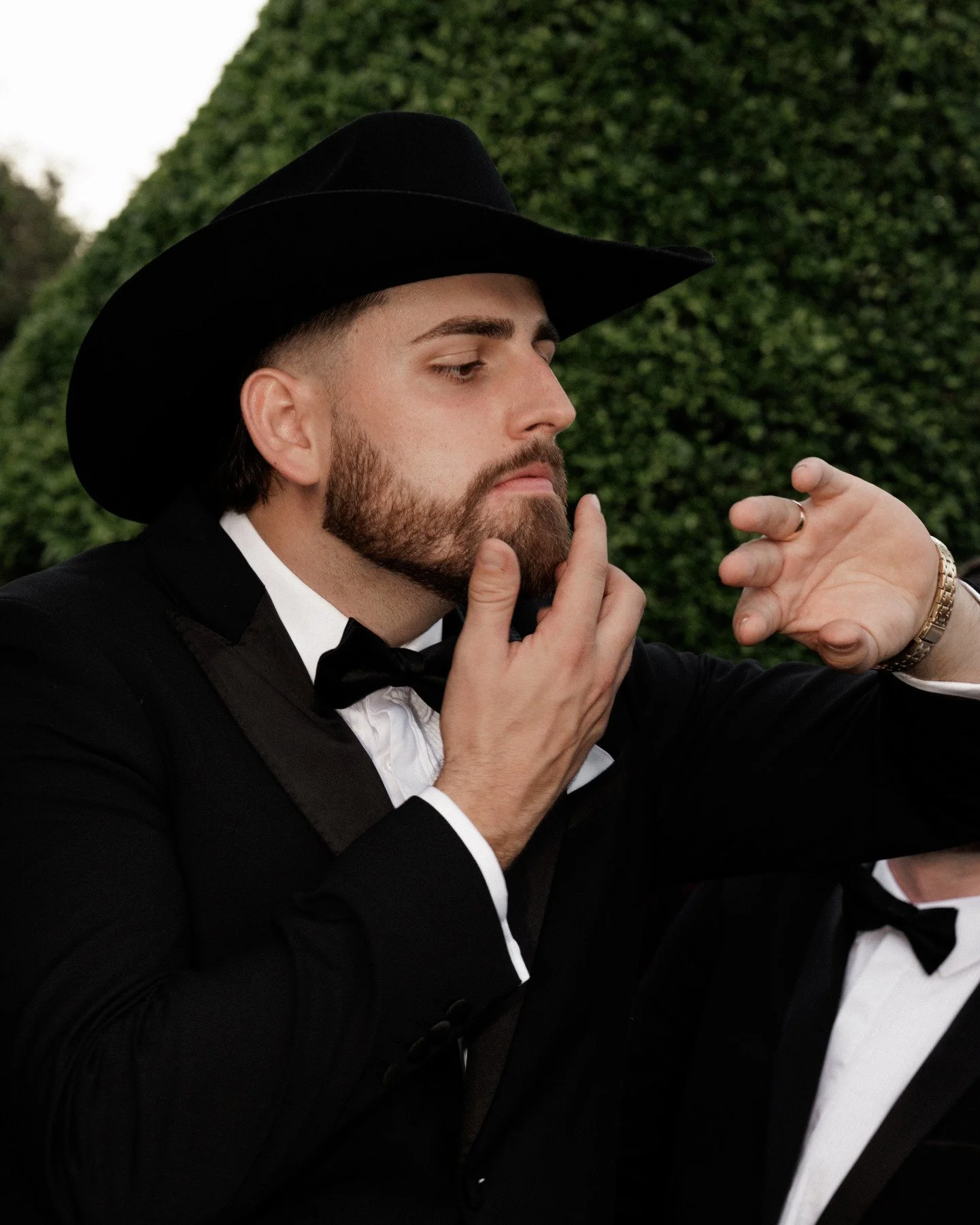 A man in a tuxedo and cowboy hat gently touching his chin, with another person's hand near his face, outdoors with a hedge in the background.