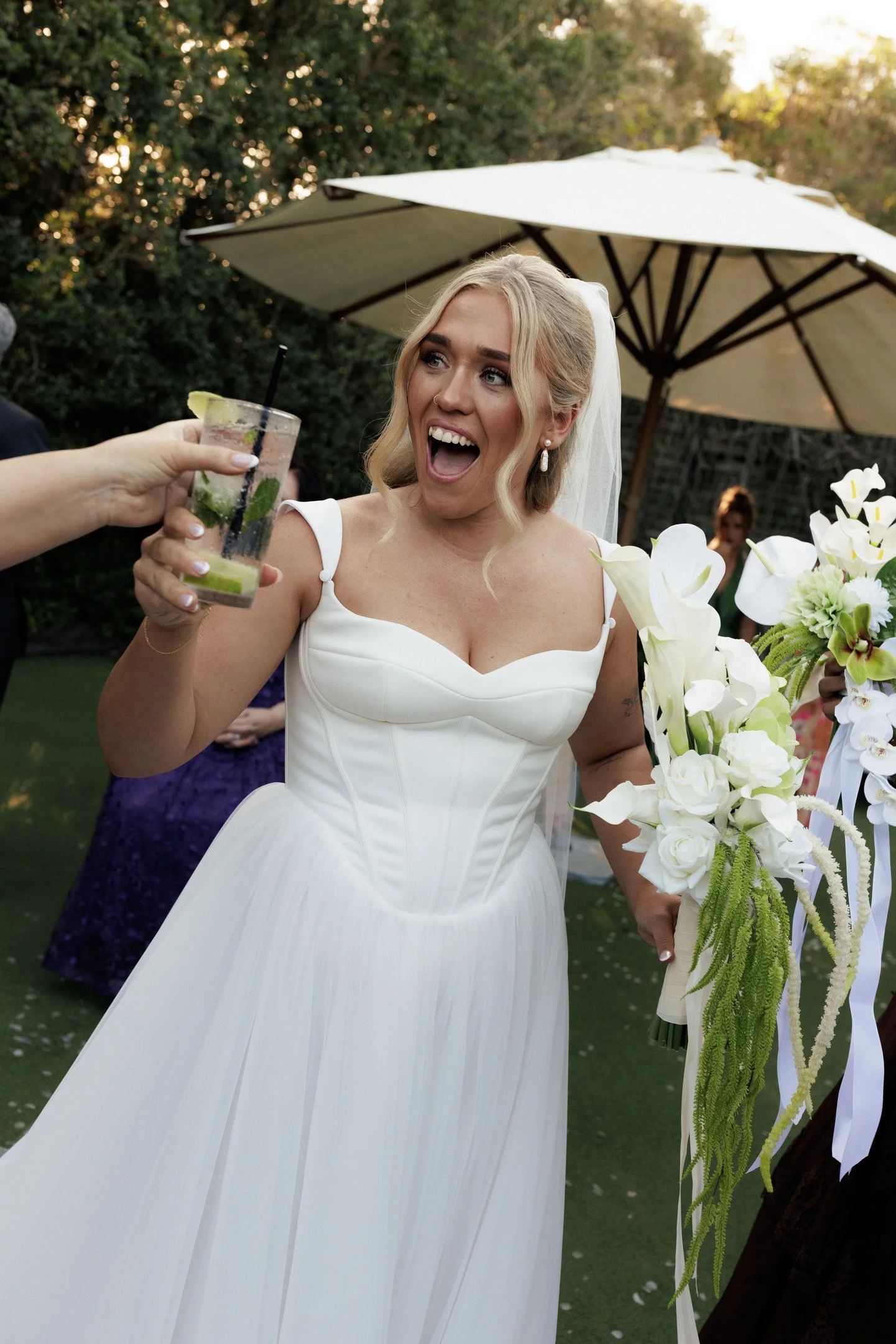 A woman in a white wedding dress is smiling with her mouth open and holding a bouquet of white flowers. She is outdoors next to a tree with large white flowers, and a person is handing her a drink with lime and mint.