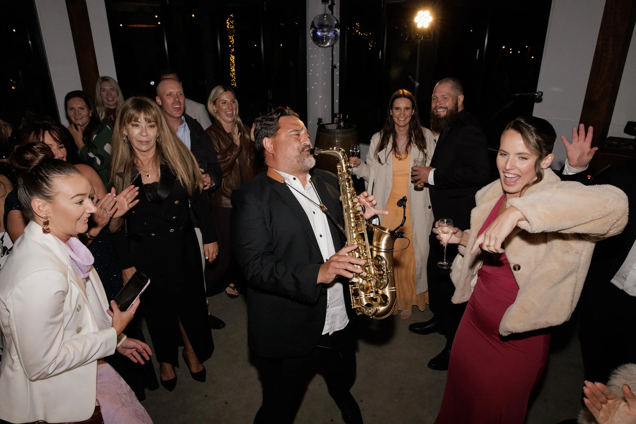A lively indoor party scene with people dancing, smiling, and enjoying drinks. A man in a tuxedo is playing the saxophone, and a woman in a red dress with a fur coat is dancing nearby. The background features large windows, evening sky, string lights