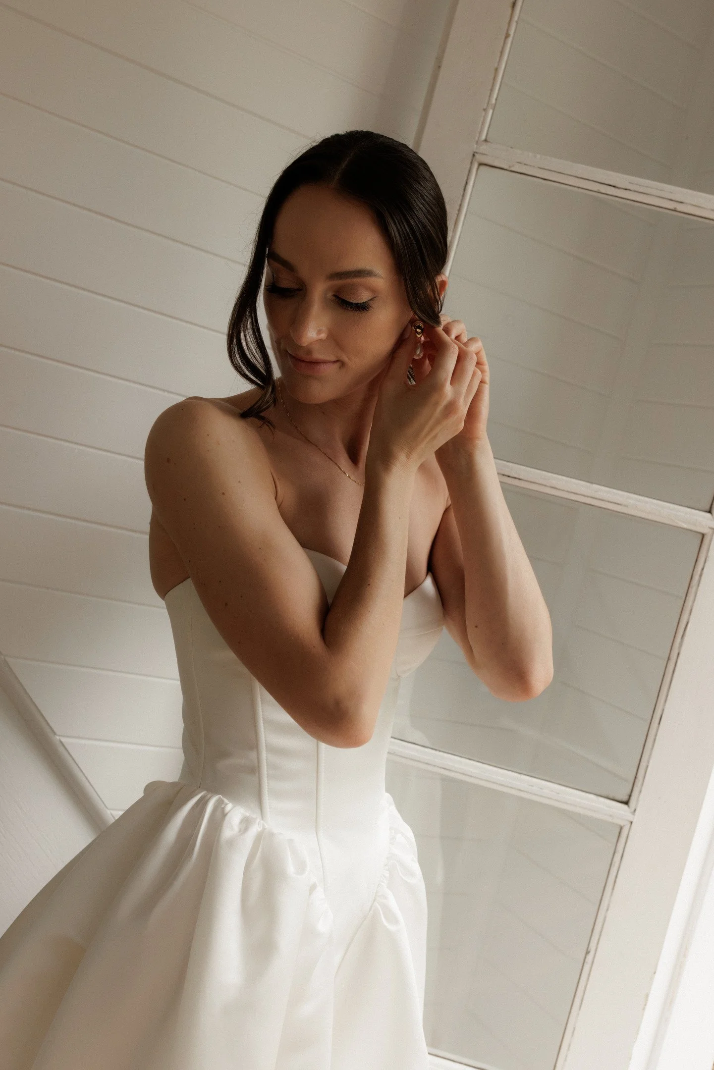 A woman with dark hair is putting on an earring in front of a mirror, wearing a strapless white dress with a skirt that has gathered fabric at the waist.
