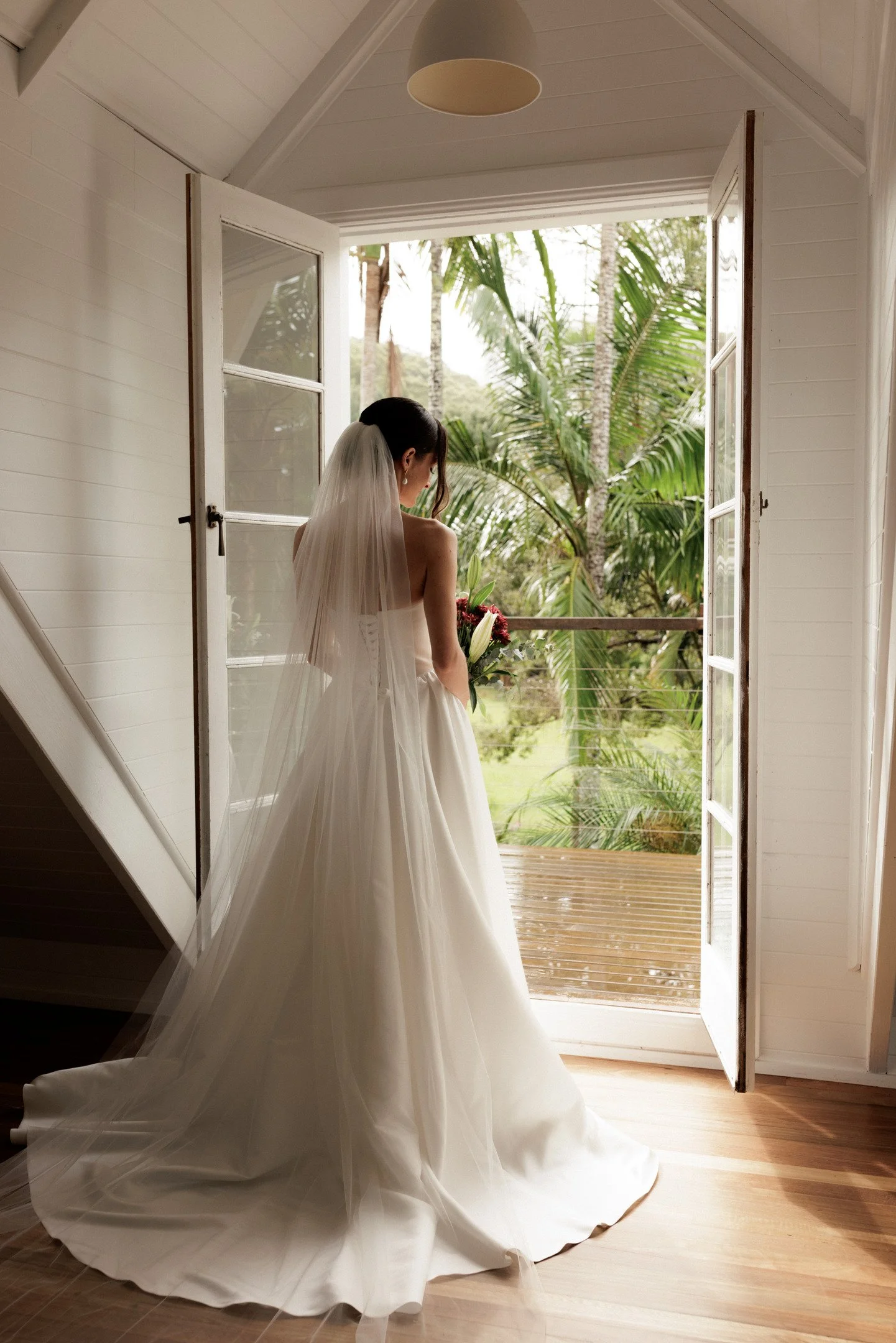 A bride standing in an open doorway with tropical palm trees visible outside, wearing a white wedding dress and veil, holding a bouquet of flowers.